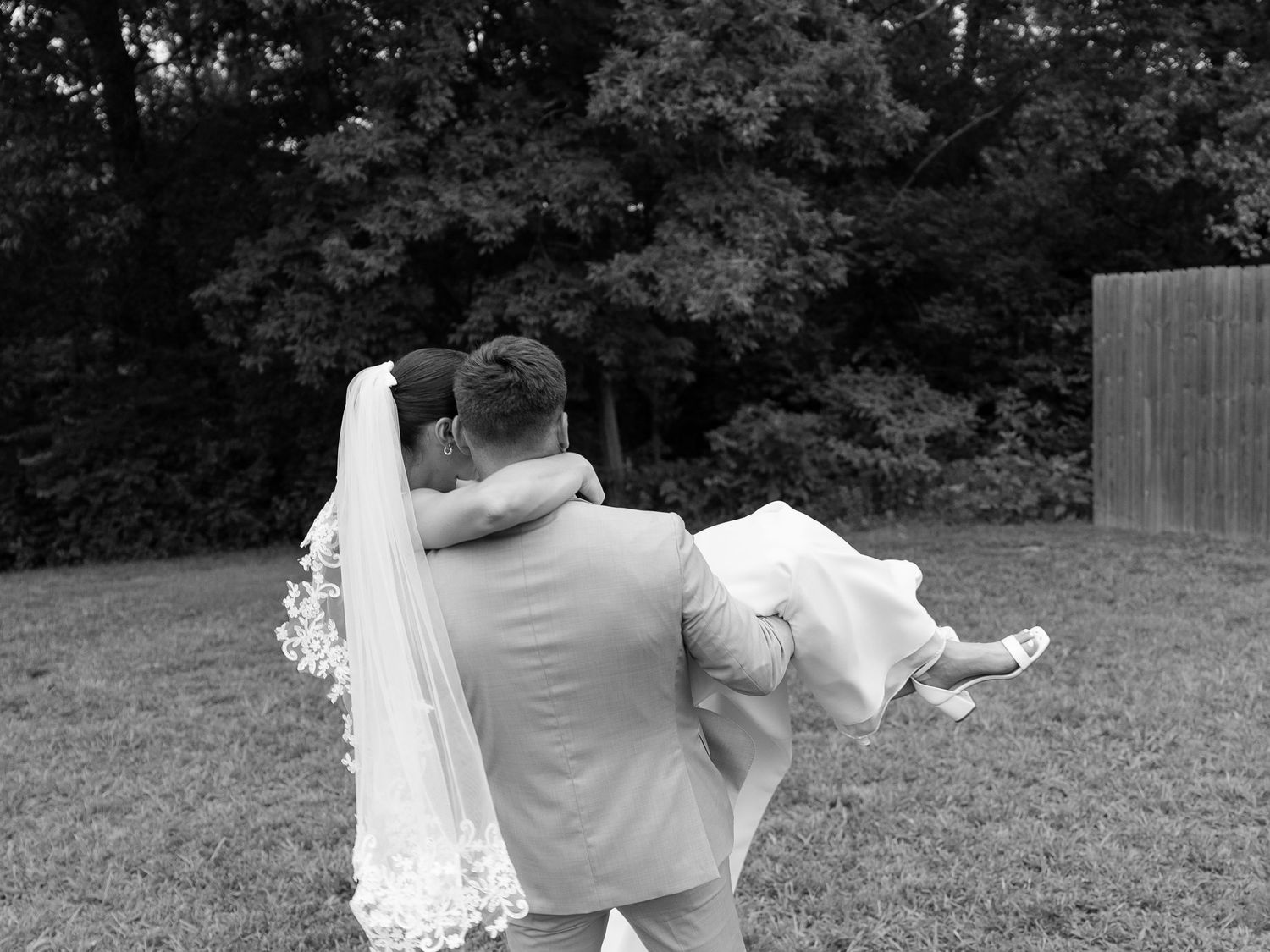 Groom carrying bride in white gown and veil across green lawn in black and white wedding photo.