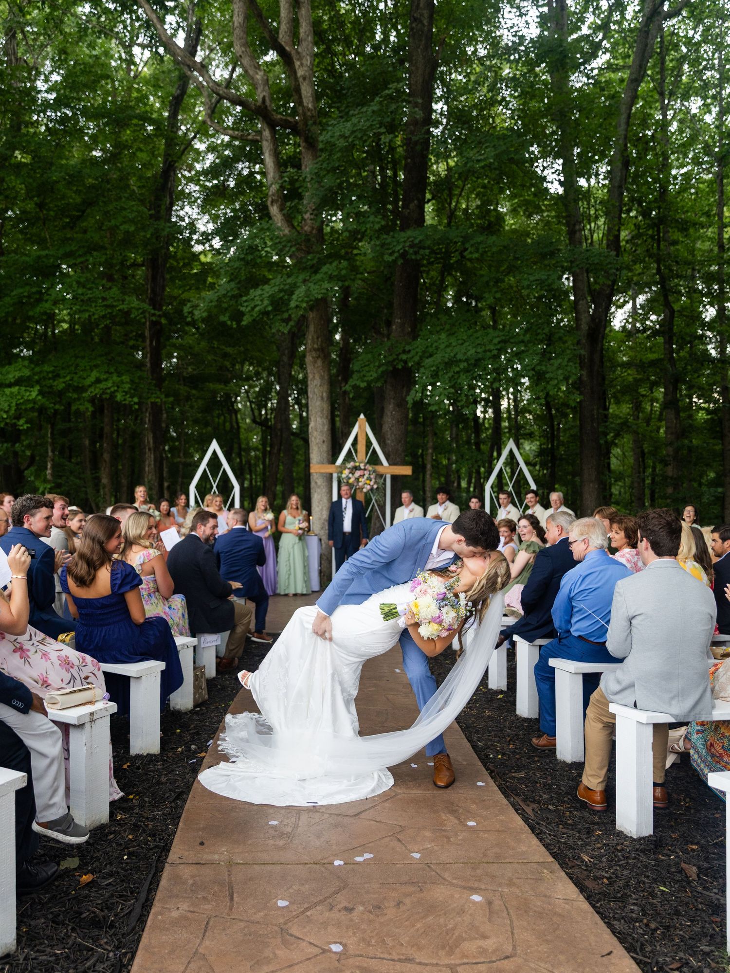 Groom dips bride for a kiss at outdoor woodland wedding ceremony with white pew seating and geometric arch backdrop.