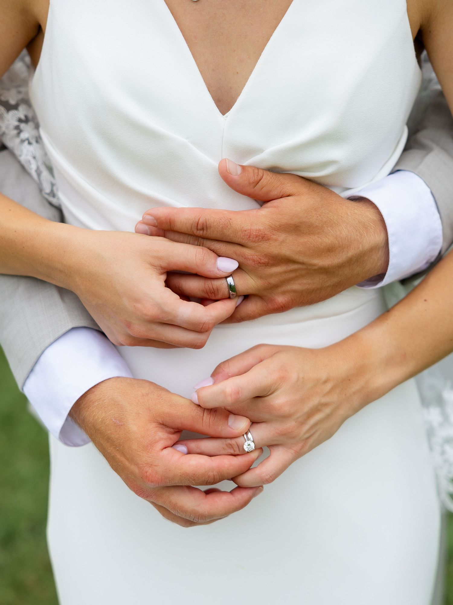Close-up of bride and groom holding hands, showing wedding rings, bride in white dress, groom in gray suit.
