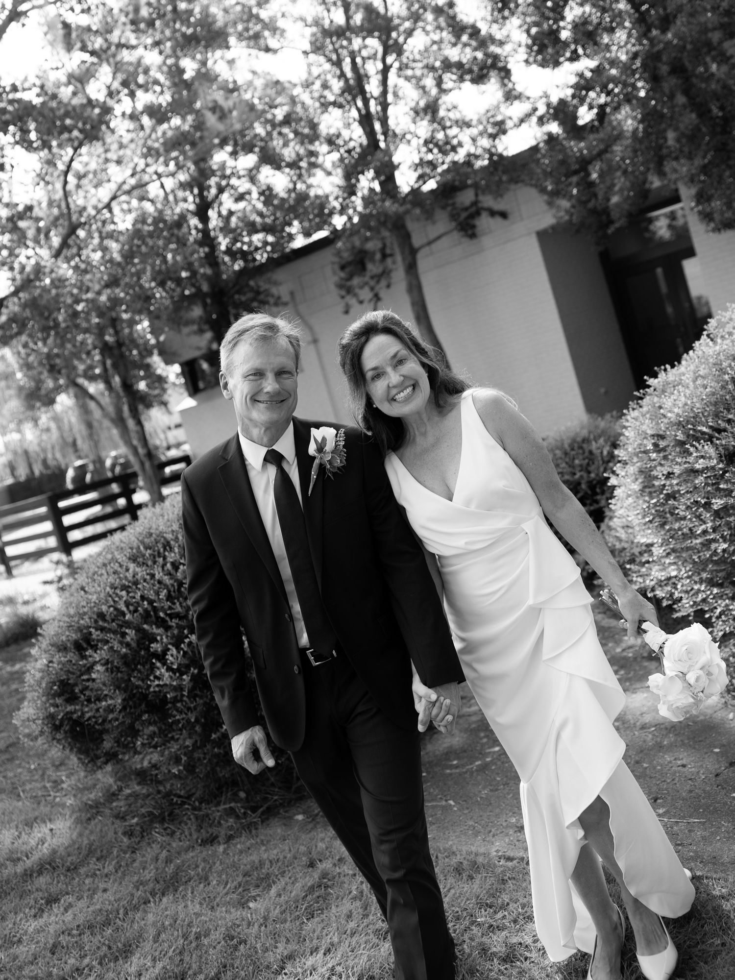 Black and white photo of a smiling couple in wedding attire walking outdoors among lush greenery.