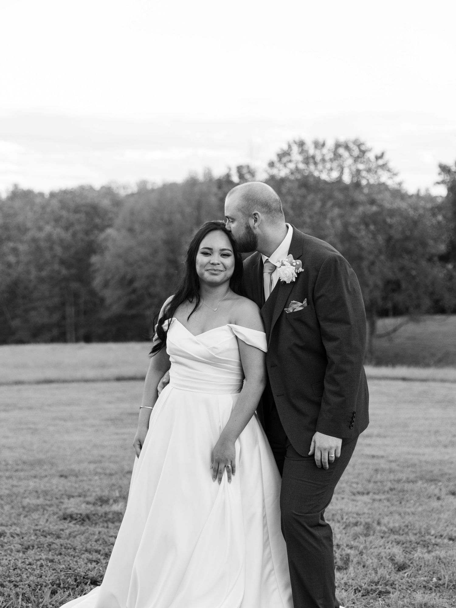 Black and white photo of a smiling bride in off-shoulder gown with groom in suit, posing in a field.