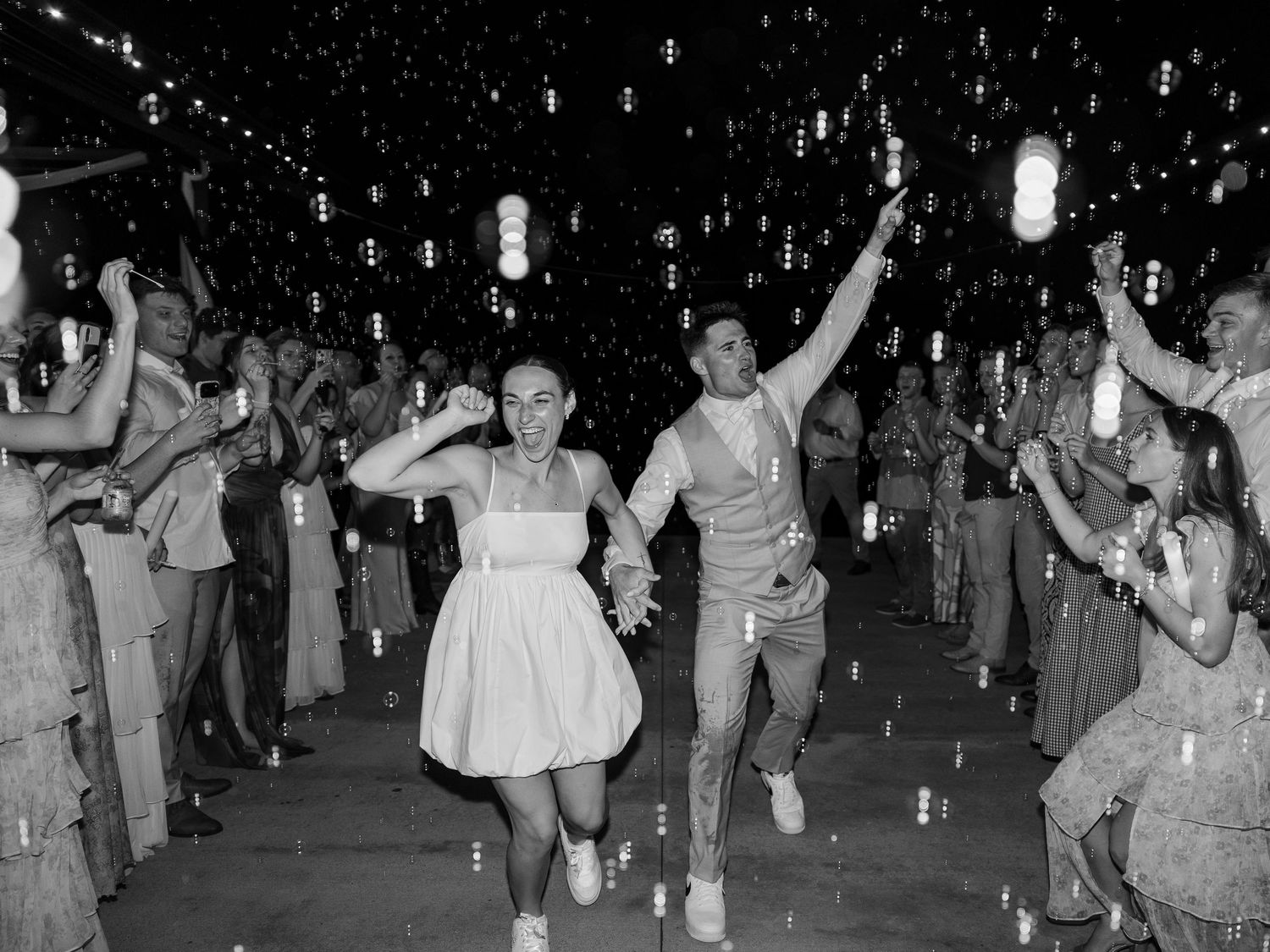 Black and white photo of a joyful couple running through a bubble-filled wedding reception exit tunnel with guests cheering.