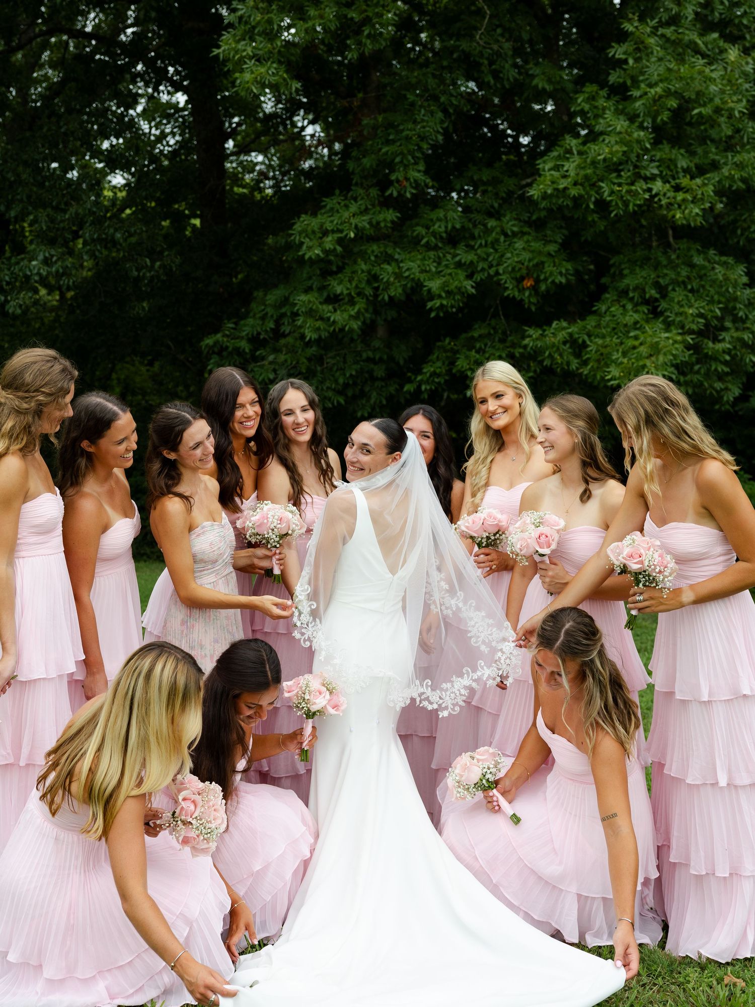 Bride in white gown and veil surrounded by bridesmaids in pink tiered dresses holding rose bouquets outdoors.