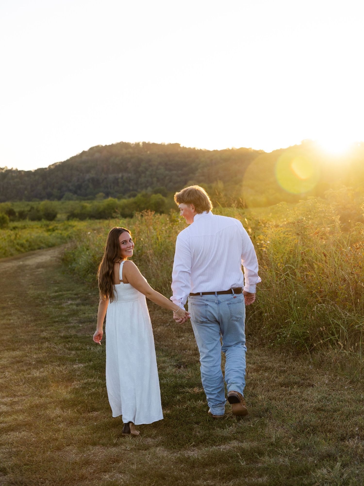 Couple holding hands walking through golden field at sunset with lush green hills in background.