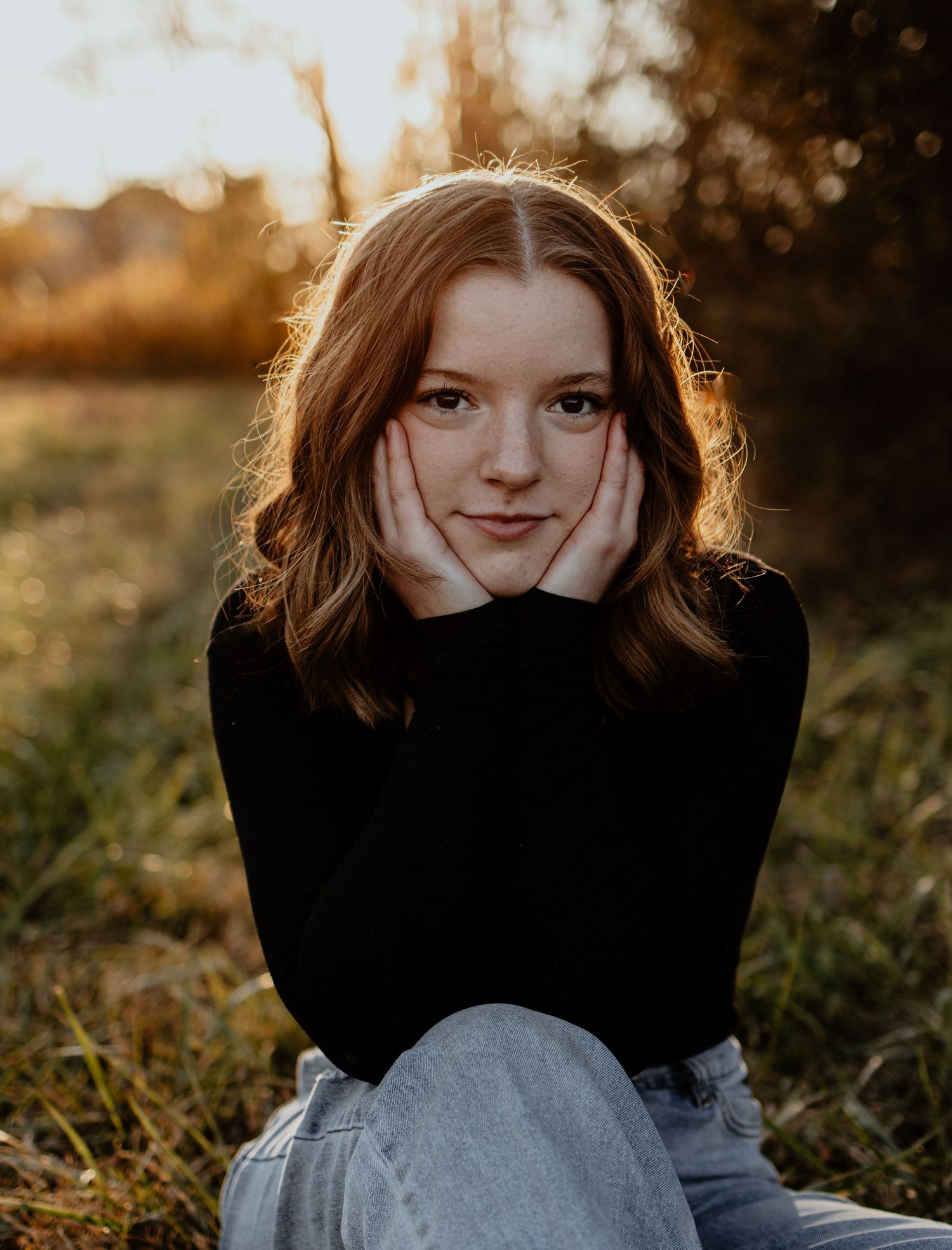 A person in a black sweater sits outdoors during golden hour, hands resting on their cheeks with sunlit hair glowing.