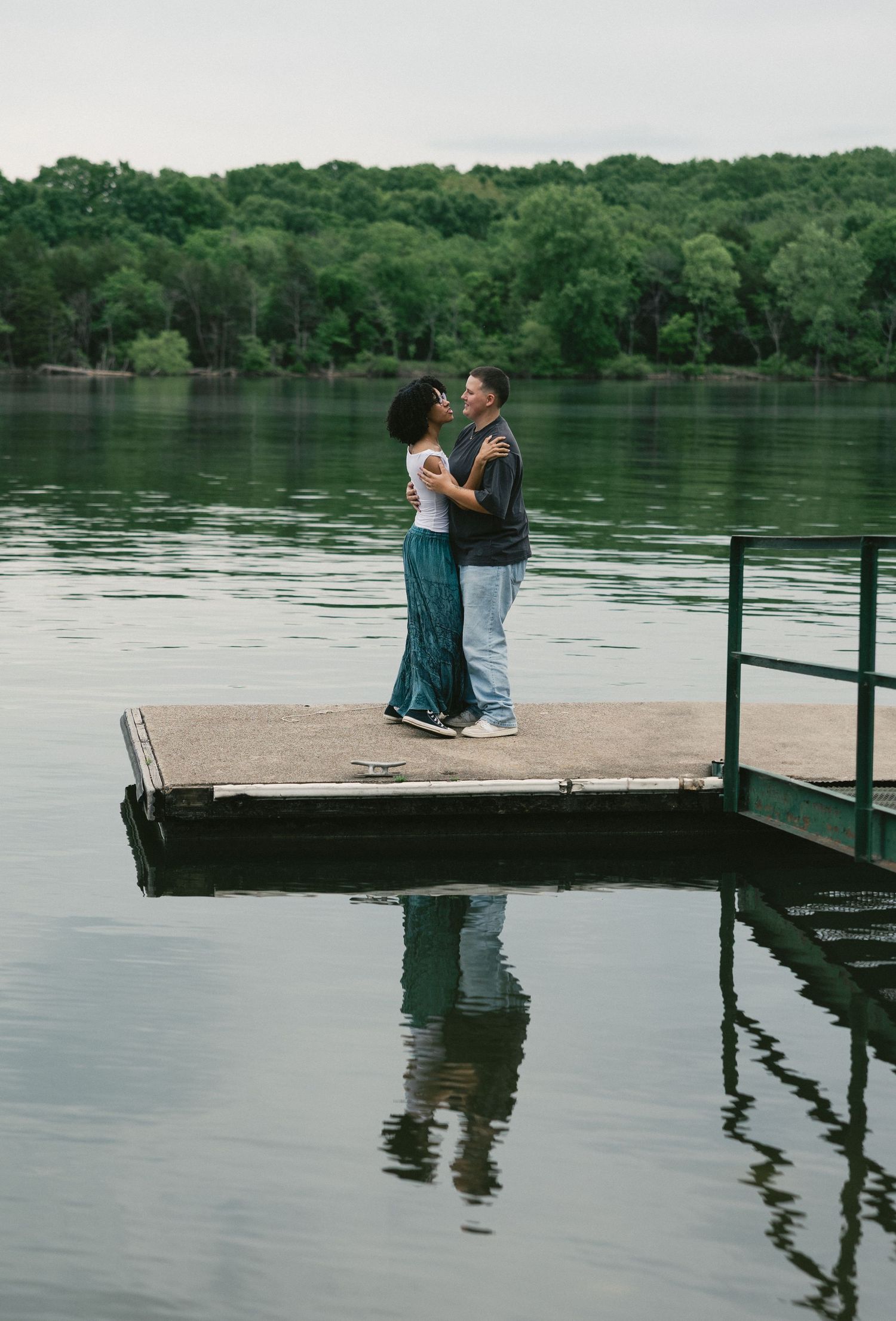 Couple sitting together on wooden dock by serene lake surrounded by lush green trees.