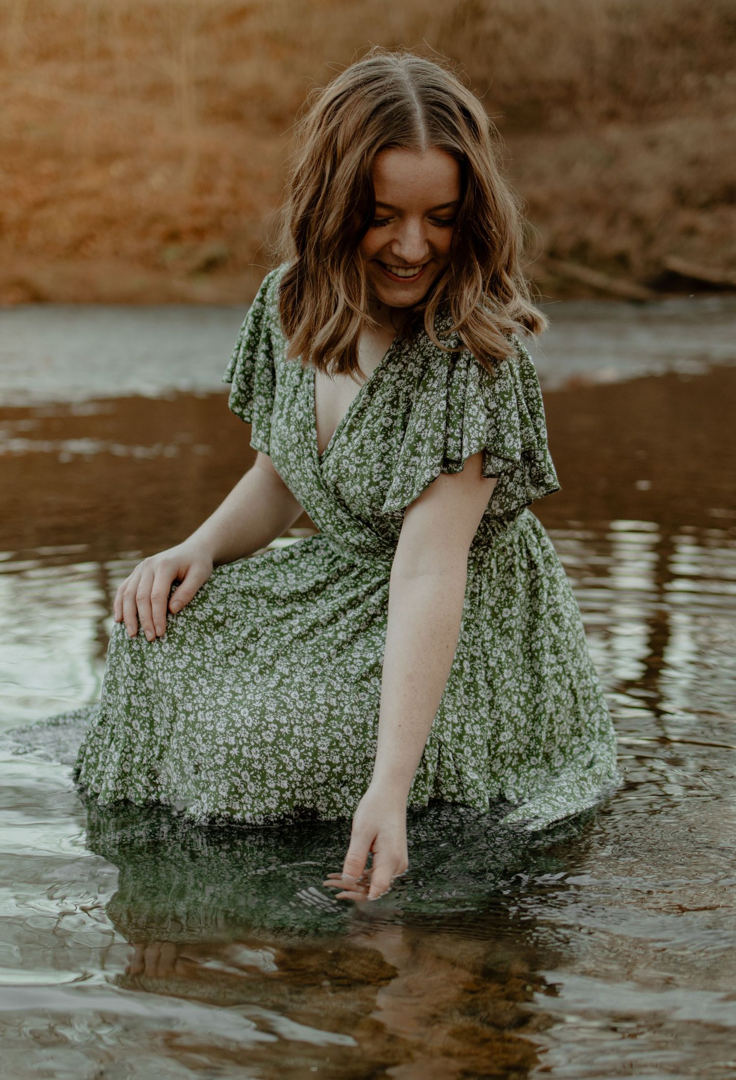 Figure in flowing green floral dress reaches into reflective water creating ripples at golden hour.