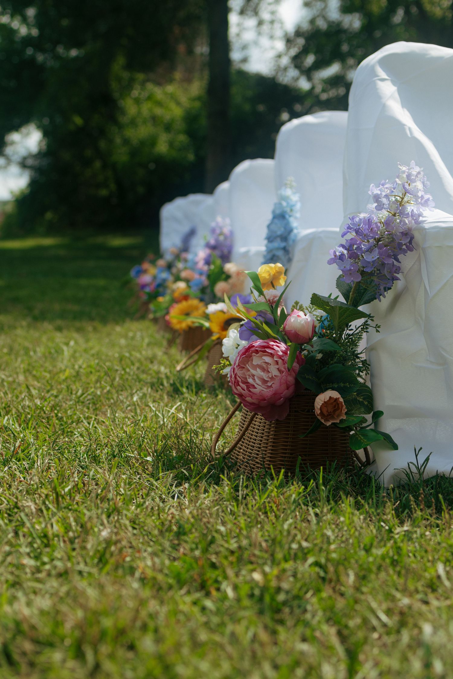 Colorful floral arrangements with peonies and wildflowers line white chairs on grass at an outdoor event setup.