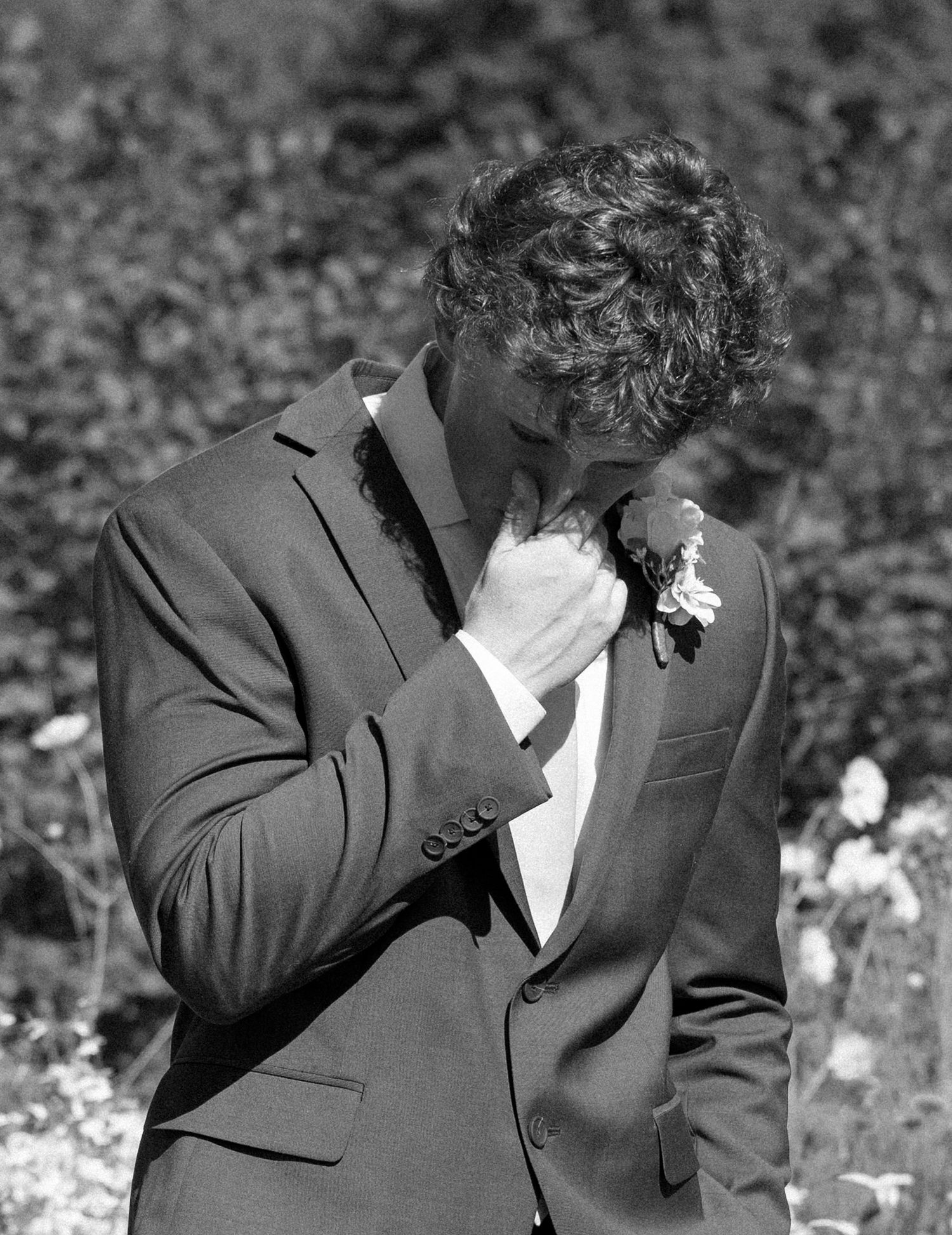 Emotional black and white photo of someone in a grey suit with a boutonniere covering their face with their hand.