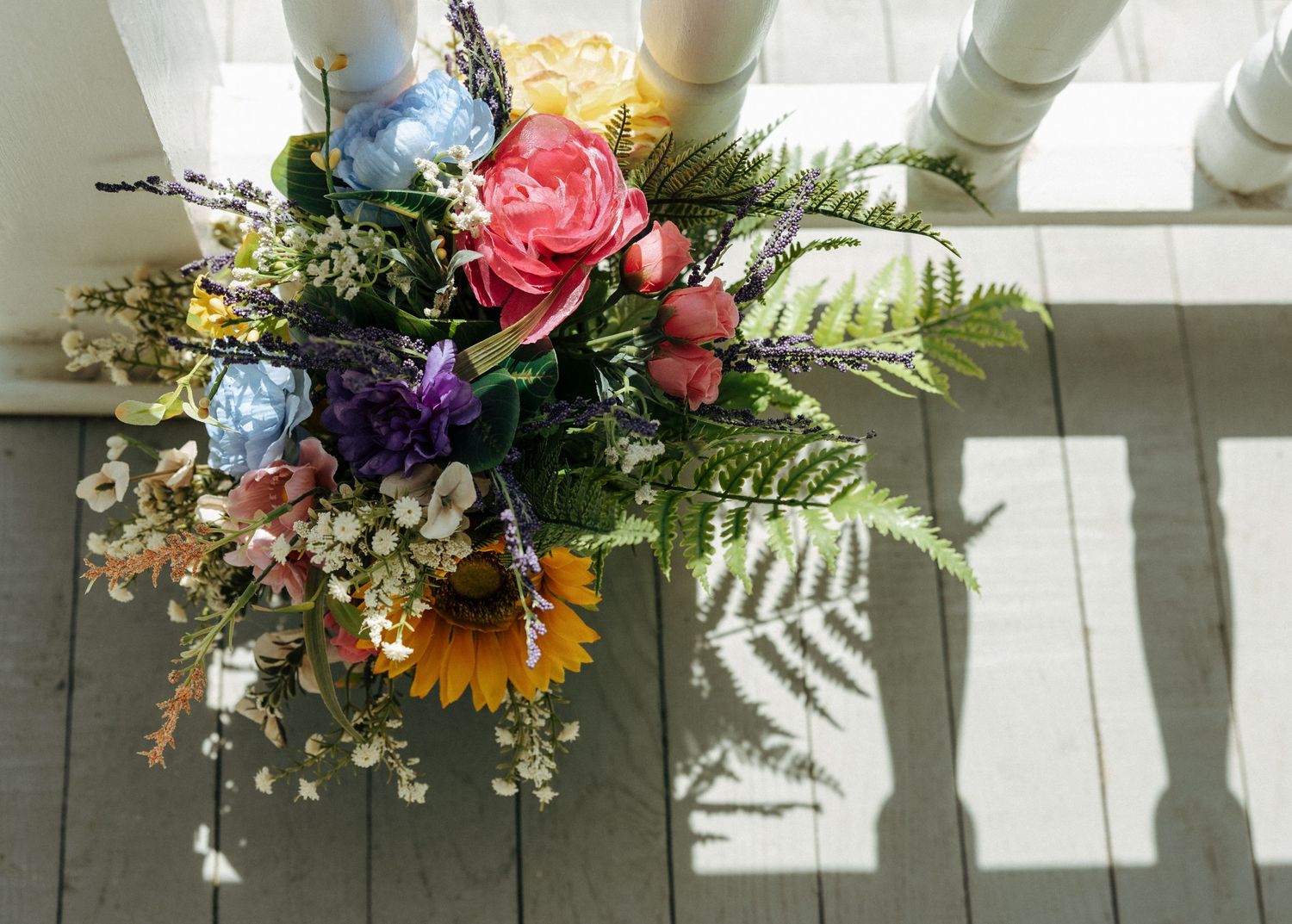 A vibrant flower arrangement with sunflowers, pink roses, blue hydrangeas, and ferns casting shadows on a white surface.
