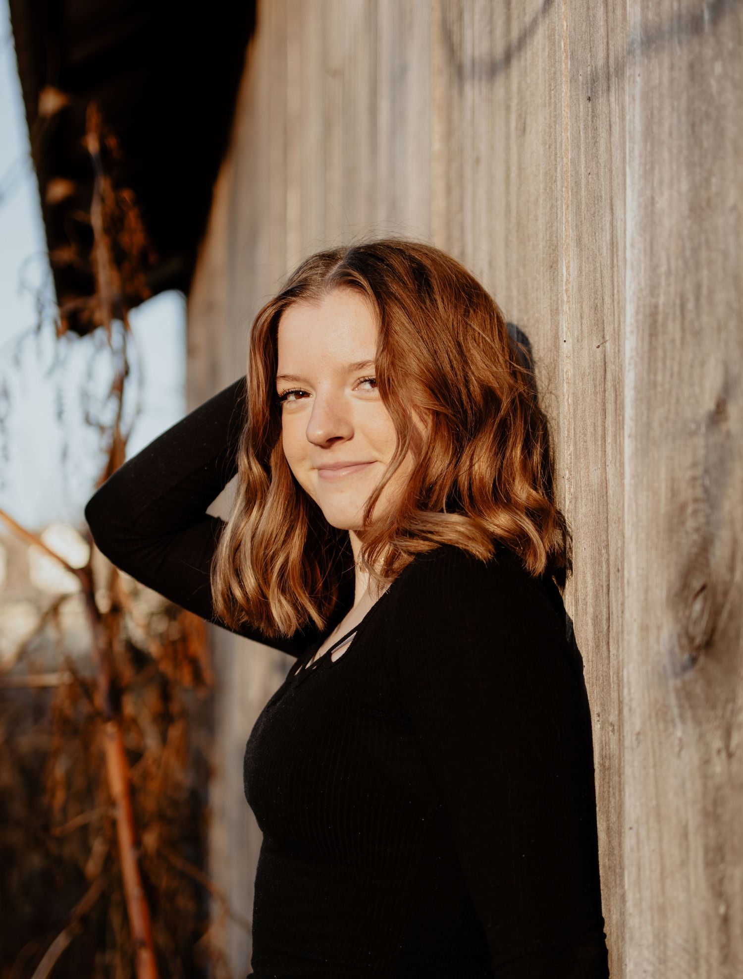 Person in a black sweater with wavy copper hair poses against a pale wall in warm sunlight.