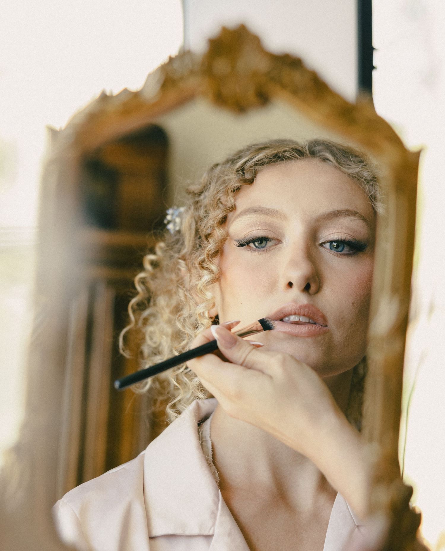 A fashion photograph reflected in an ornate mirror shows someone applying makeup with a brush.