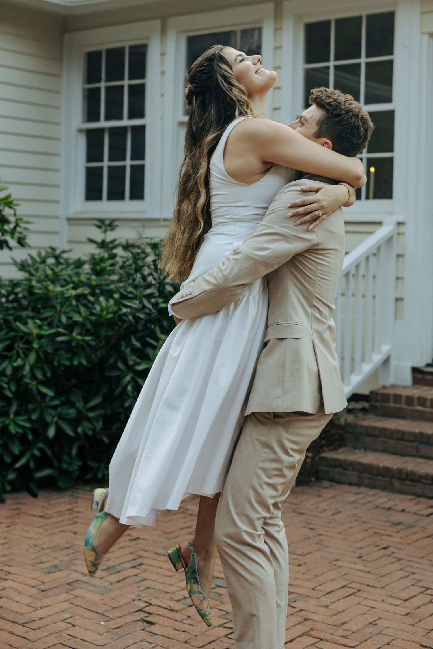 A couple embraces joyfully on a brick patio, with the bride in a white dress being lifted by her partner in tan suit.