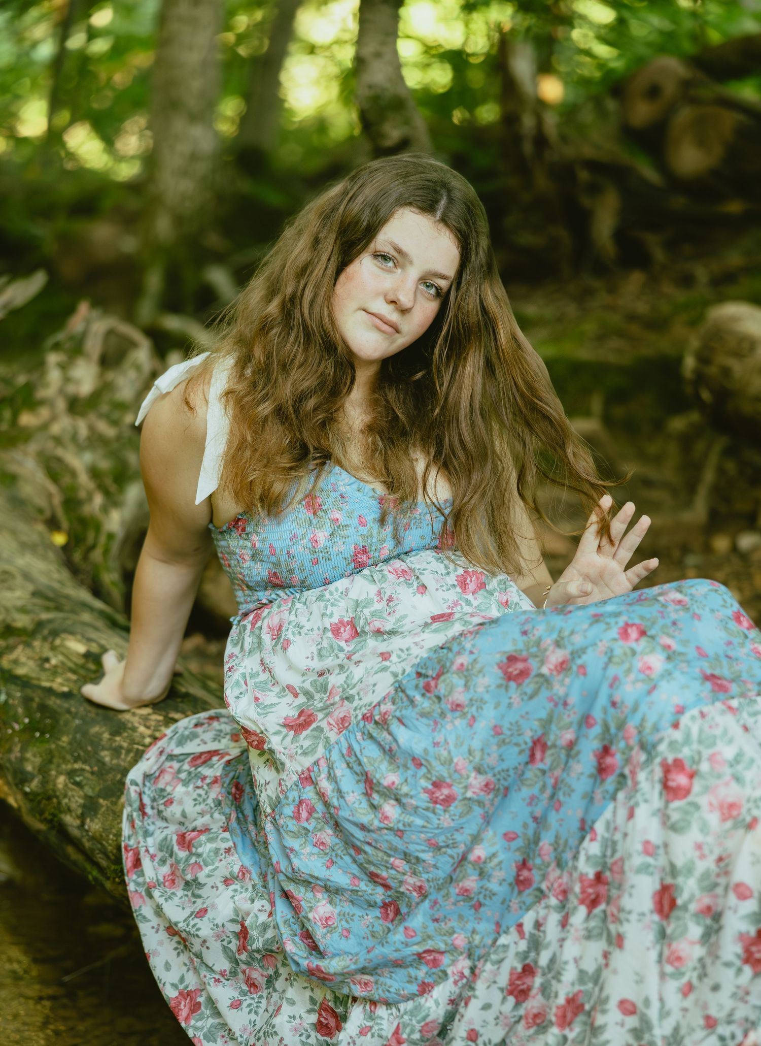 Young woman in floral blue dress poses gracefully against natural woodland background in summer photoshoot.