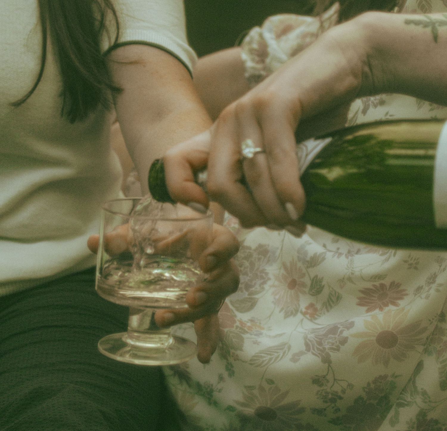 Hands pour champagne into a vintage coupe glass against a floral patterned fabric background.