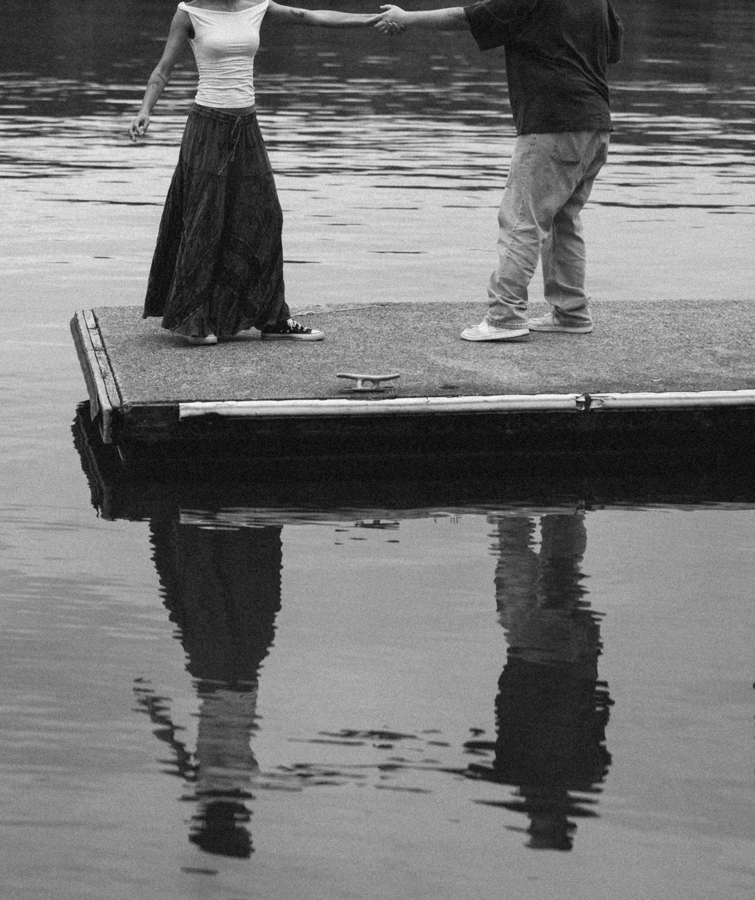 Two figures standing on a wooden dock over calm water, creating reflective silhouettes in black and white.