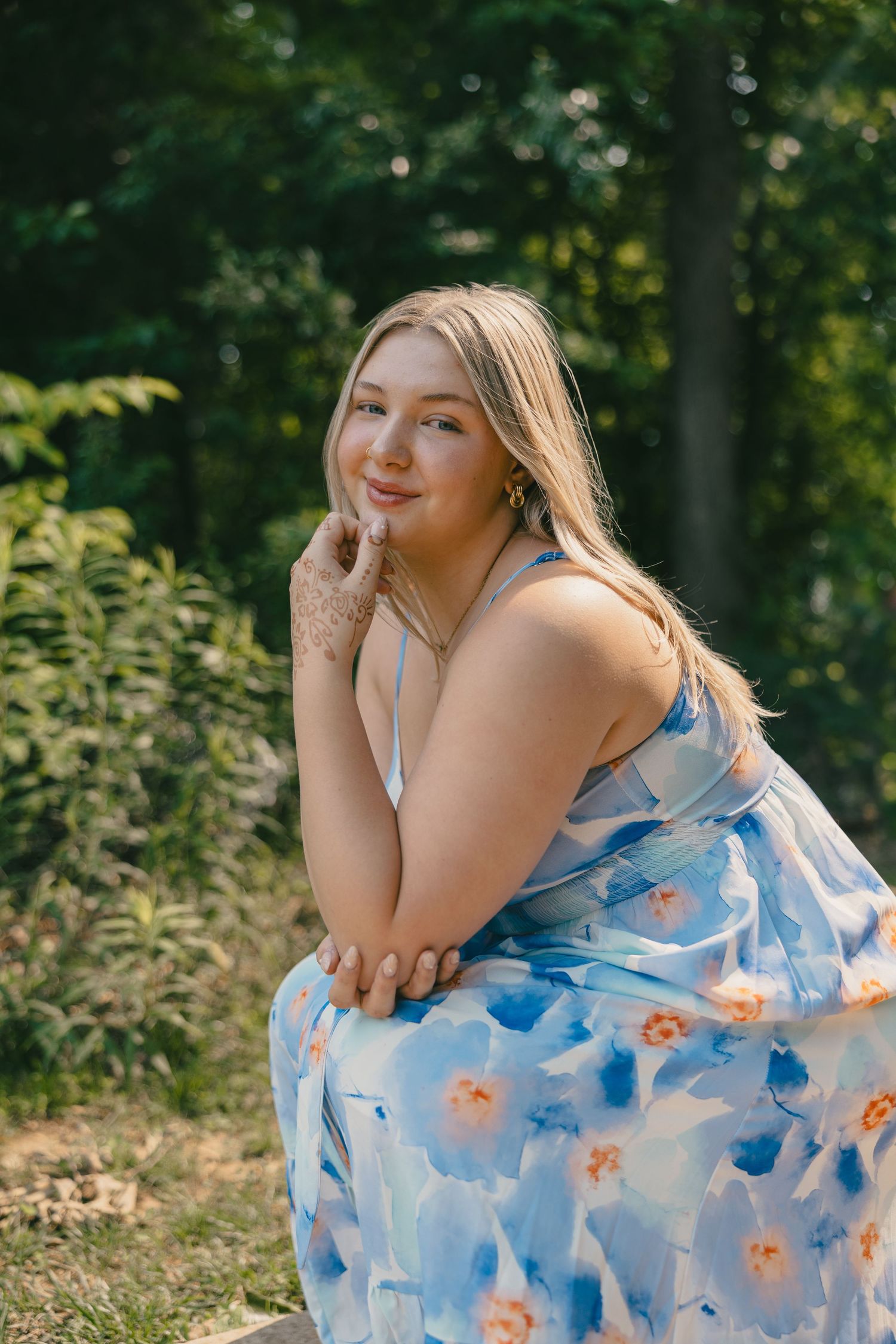 A person in a flowing blue floral dress poses outdoors in a natural setting with sunlit trees in the background.