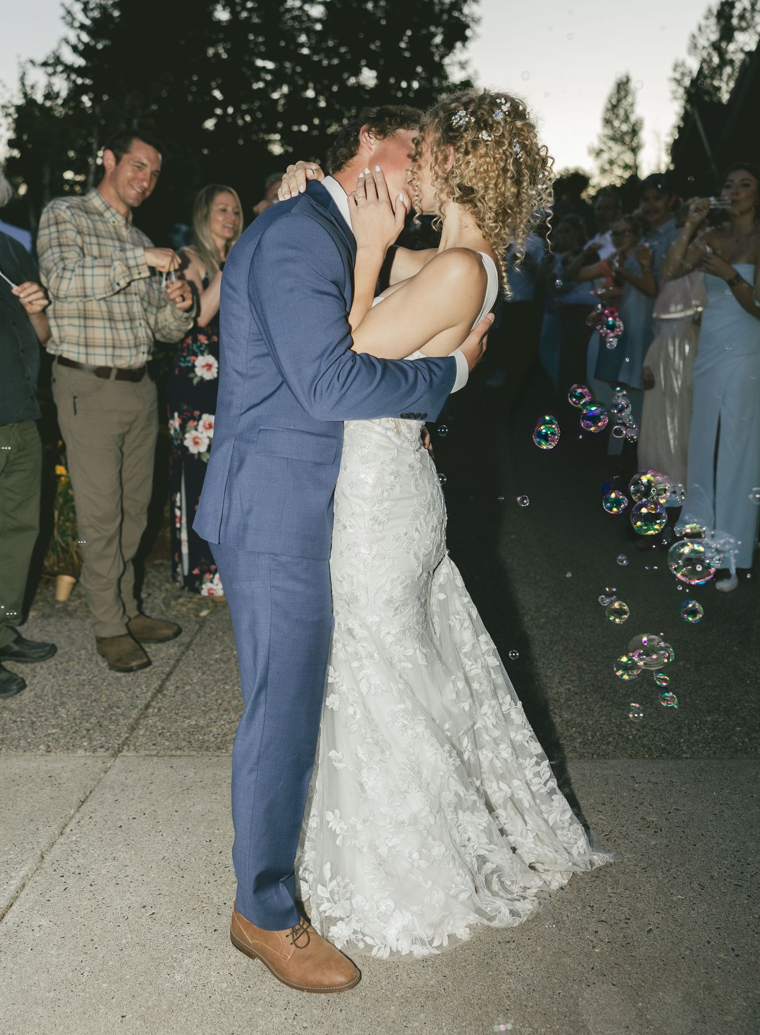 Newlywed couple shares romantic kiss surrounded by floating bubbles at outdoor evening wedding celebration.
