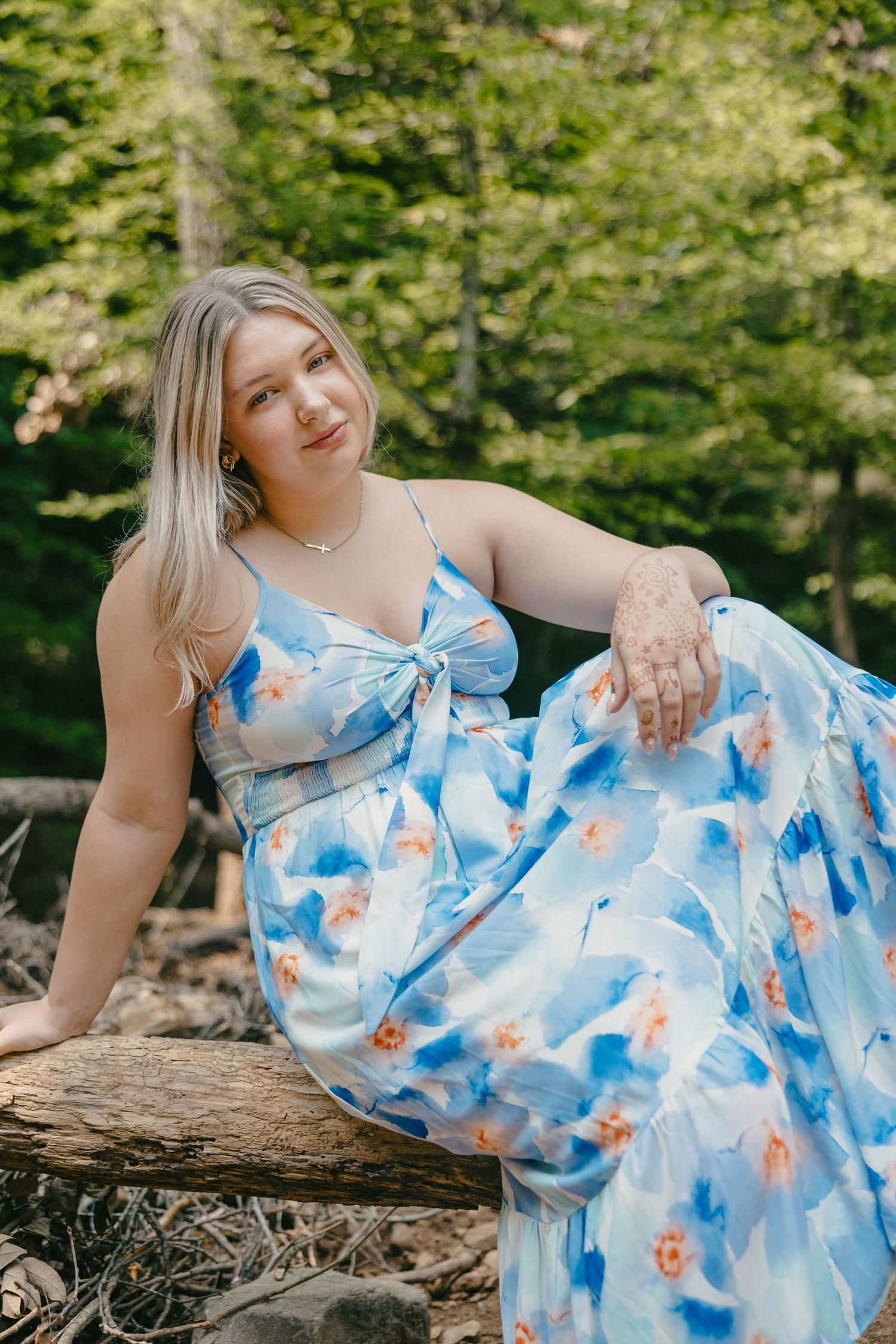 Someone wearing a flowy blue and white watercolor dress poses on a fallen tree trunk in a sunny forest setting.