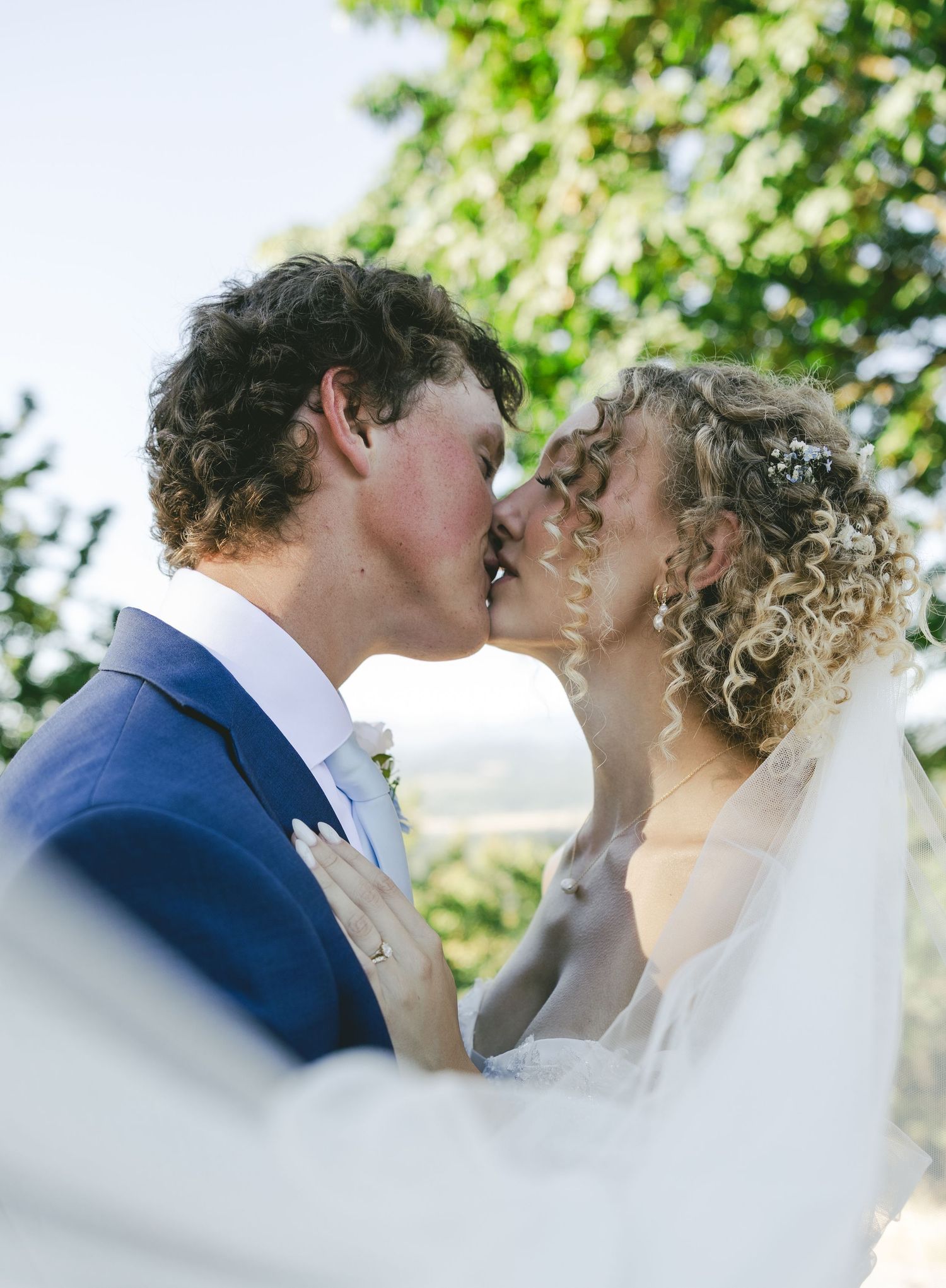 A romantic wedding kiss captured outdoors with soft natural lighting and a flowing white veil in the foreground.
