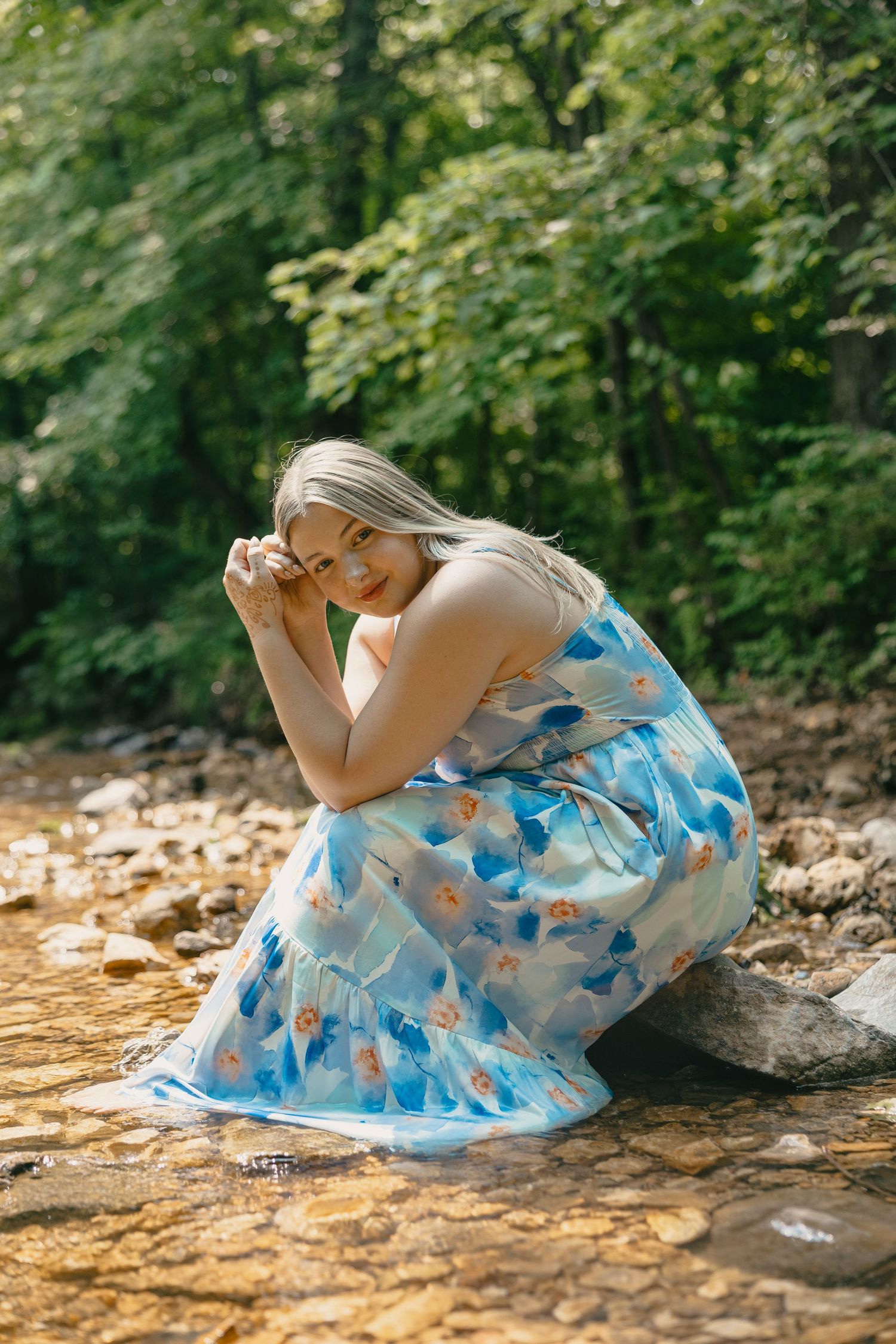 Someone wearing a blue floral dress sits on rocky terrain beside a stream in a forest setting.