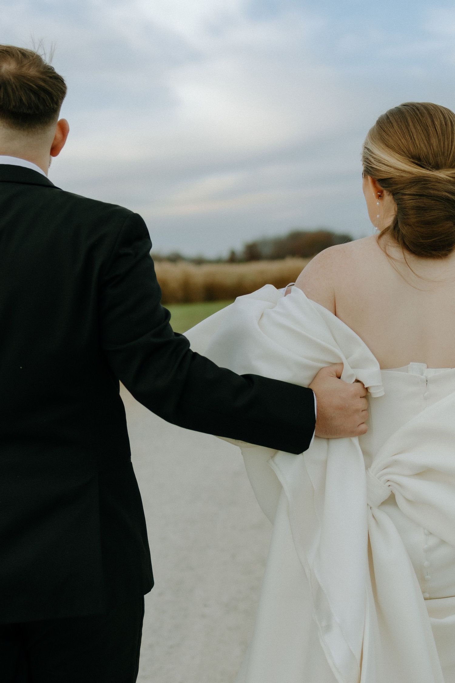 A couple in wedding attire walking together on a path with the groom helping to hold the bride's white dress.