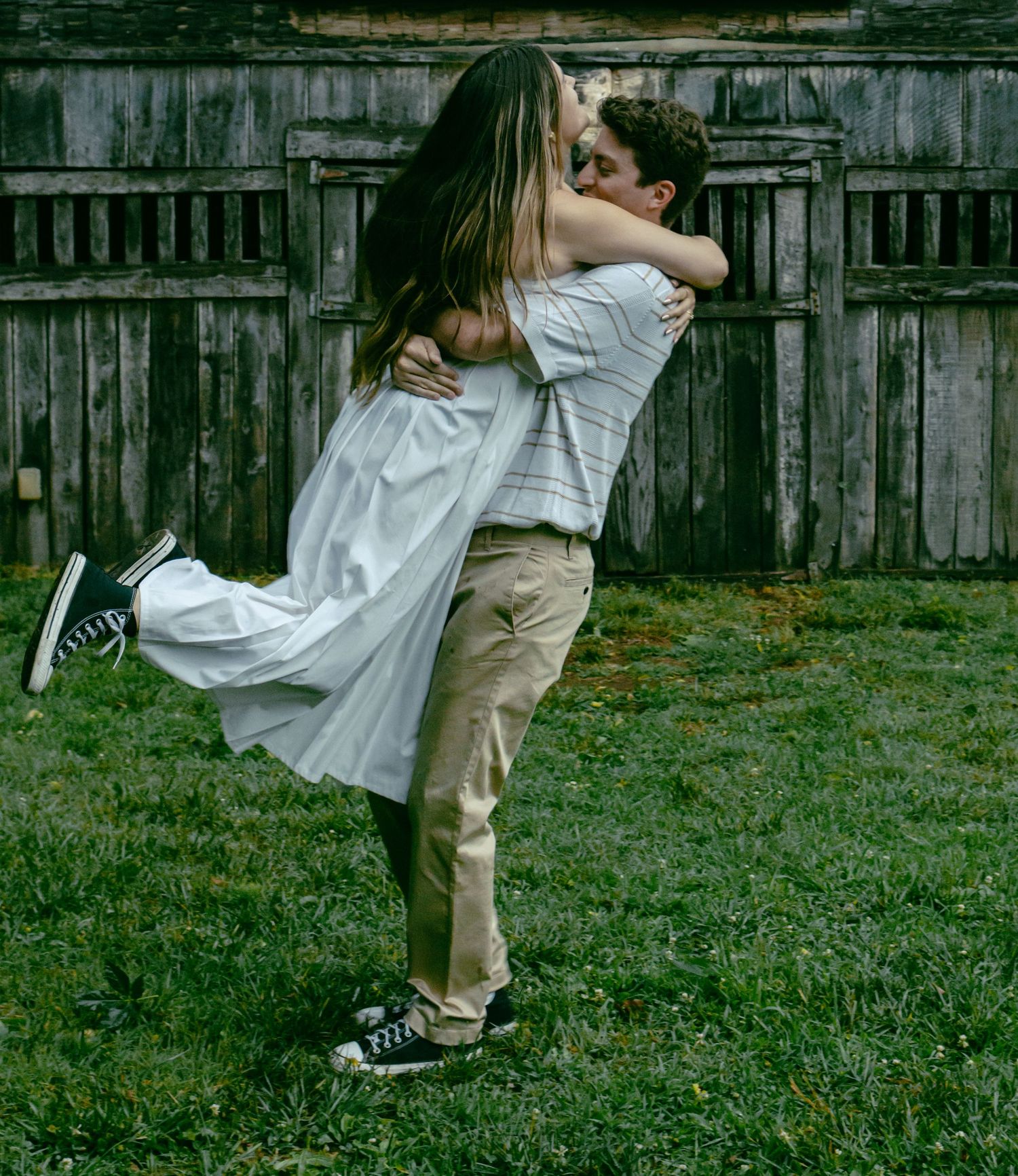 A couple embraces in an intimate moment against a rustic wooden fence backdrop while wearing casual attire.