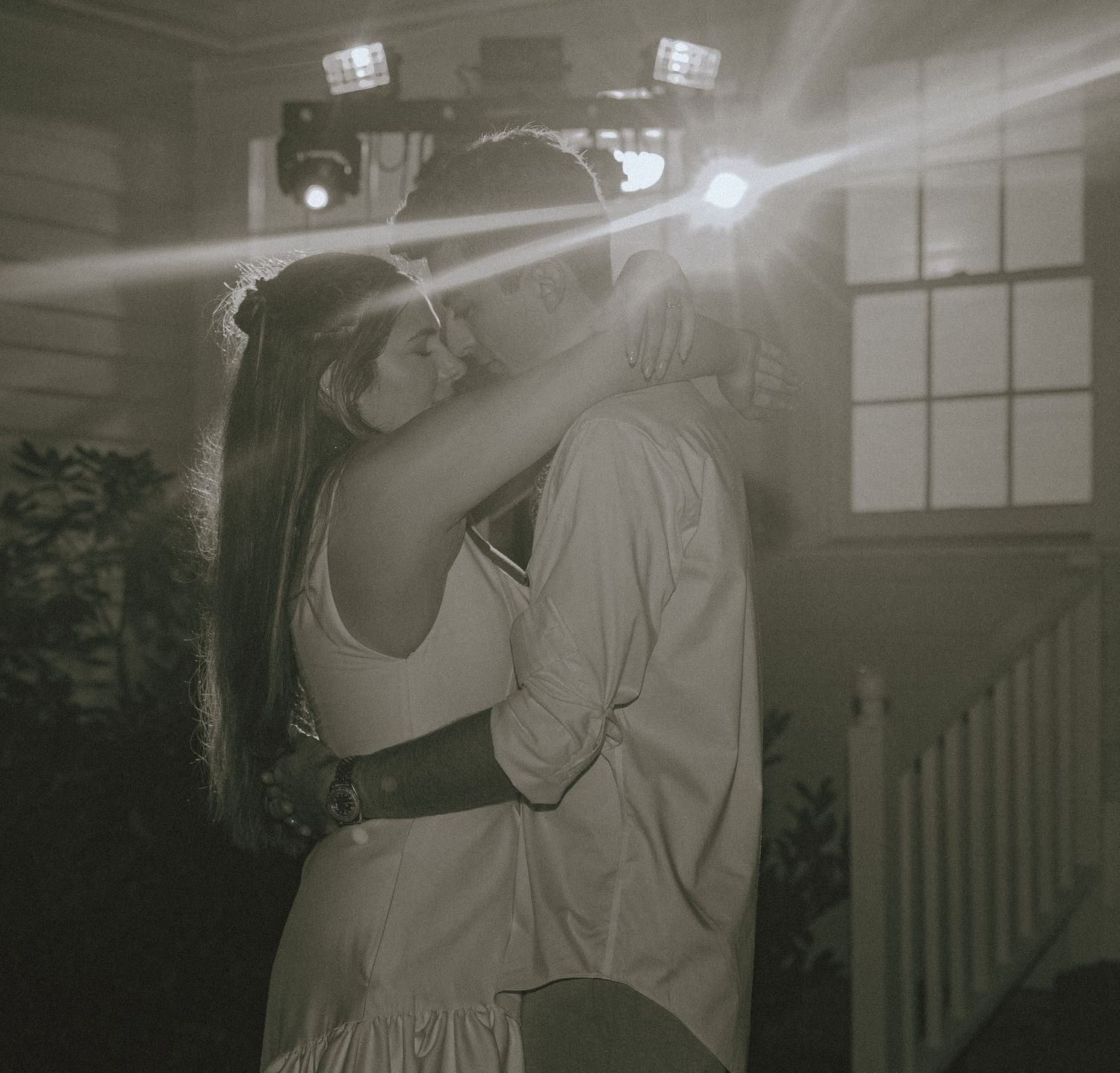 A romantic black and white photo of a couple dancing in dim lighting with streaming light beams in the background.