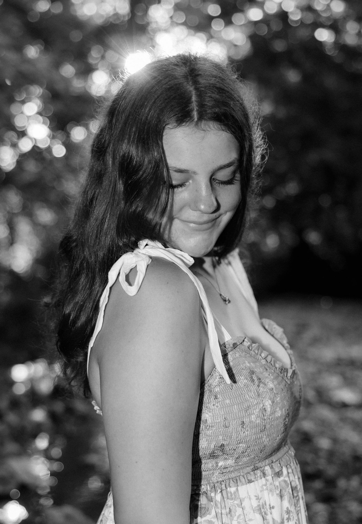 A moody black and white portrait of a young person in a summer dress standing against a bokeh forest background.