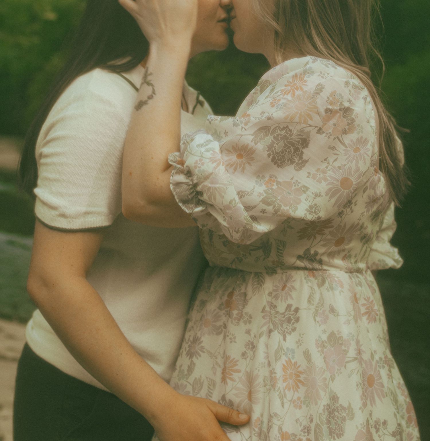 Two figures embrace in a romantic vintage-style moment wearing light-colored dresses against a blurred green background.