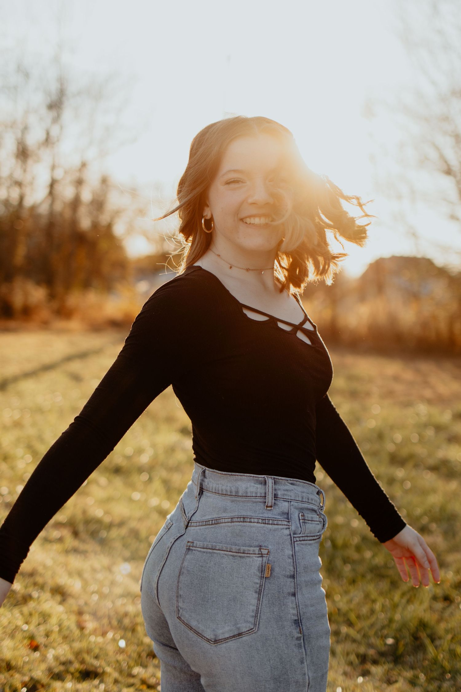 Figure wearing black long sleeve top and high waist jeans spins joyfully in golden sunlight during sunset.