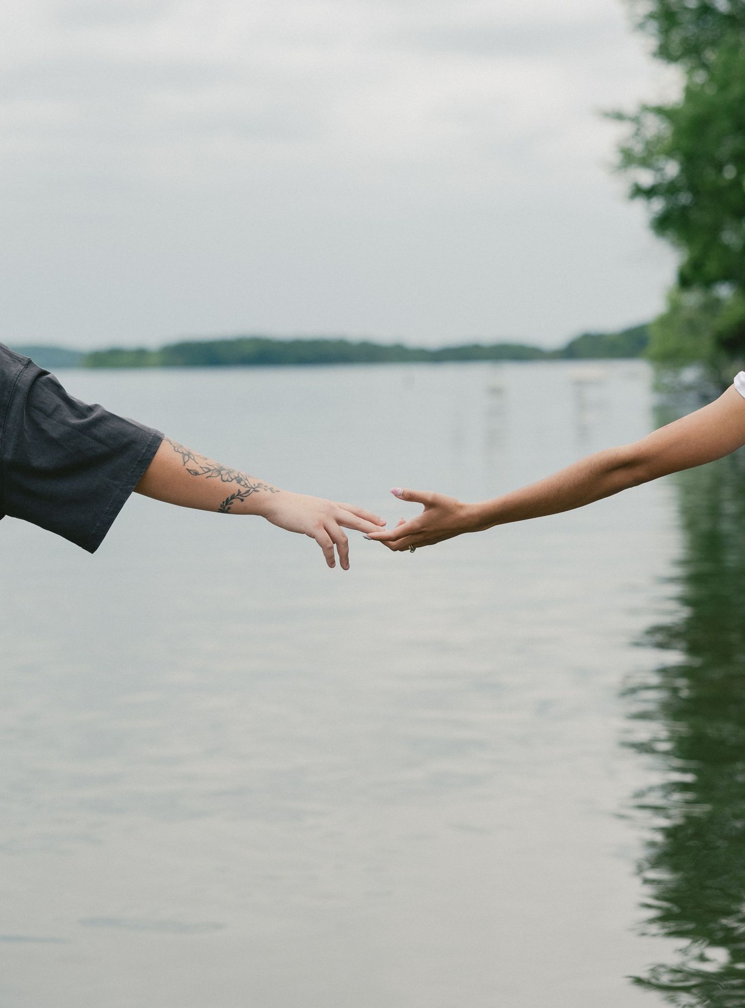 Two hands reaching towards each other against a serene lake background with trees along the shoreline.
