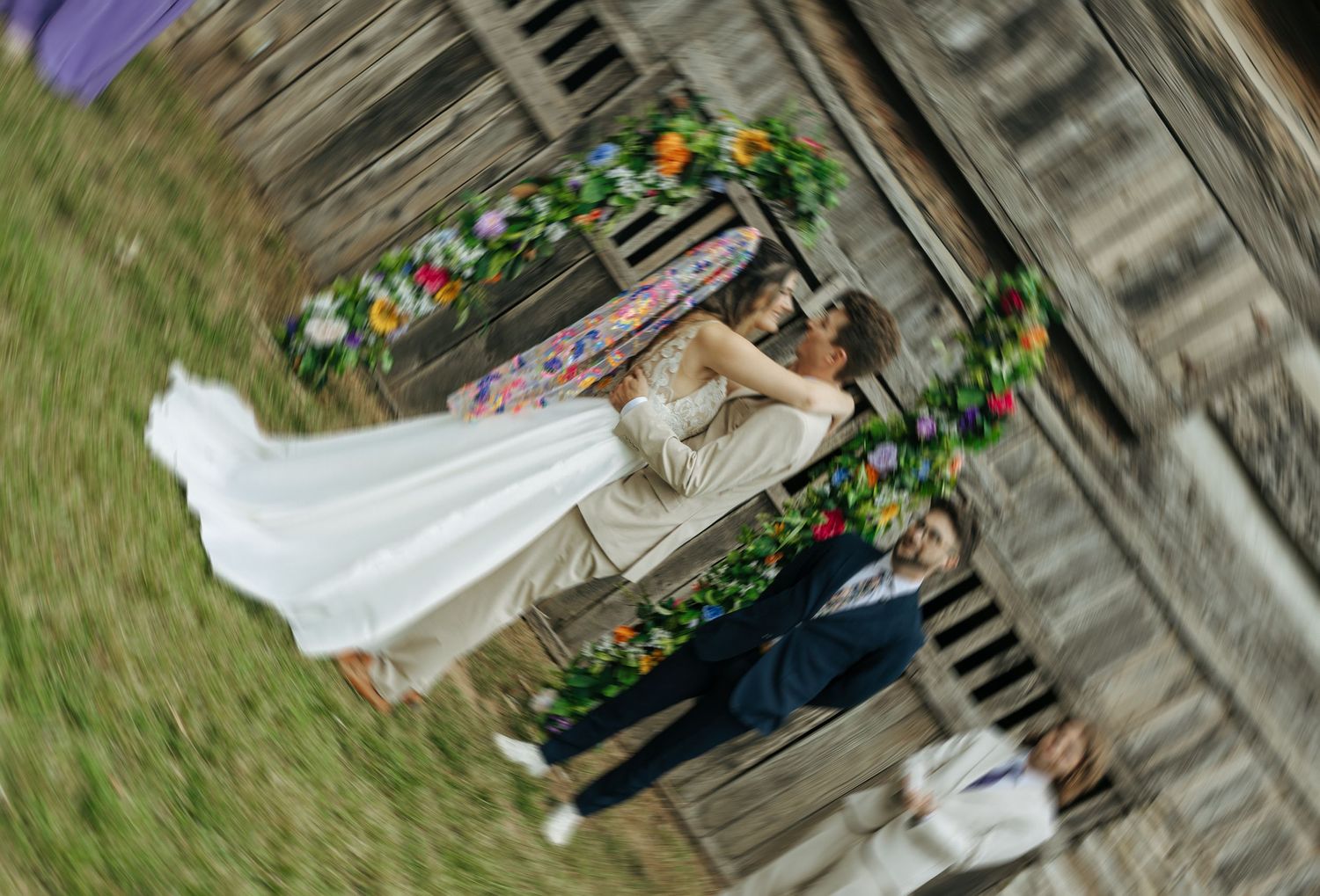 A couple embraces under a floral arch on wooden stairs in a romantic rustic outdoor setting.