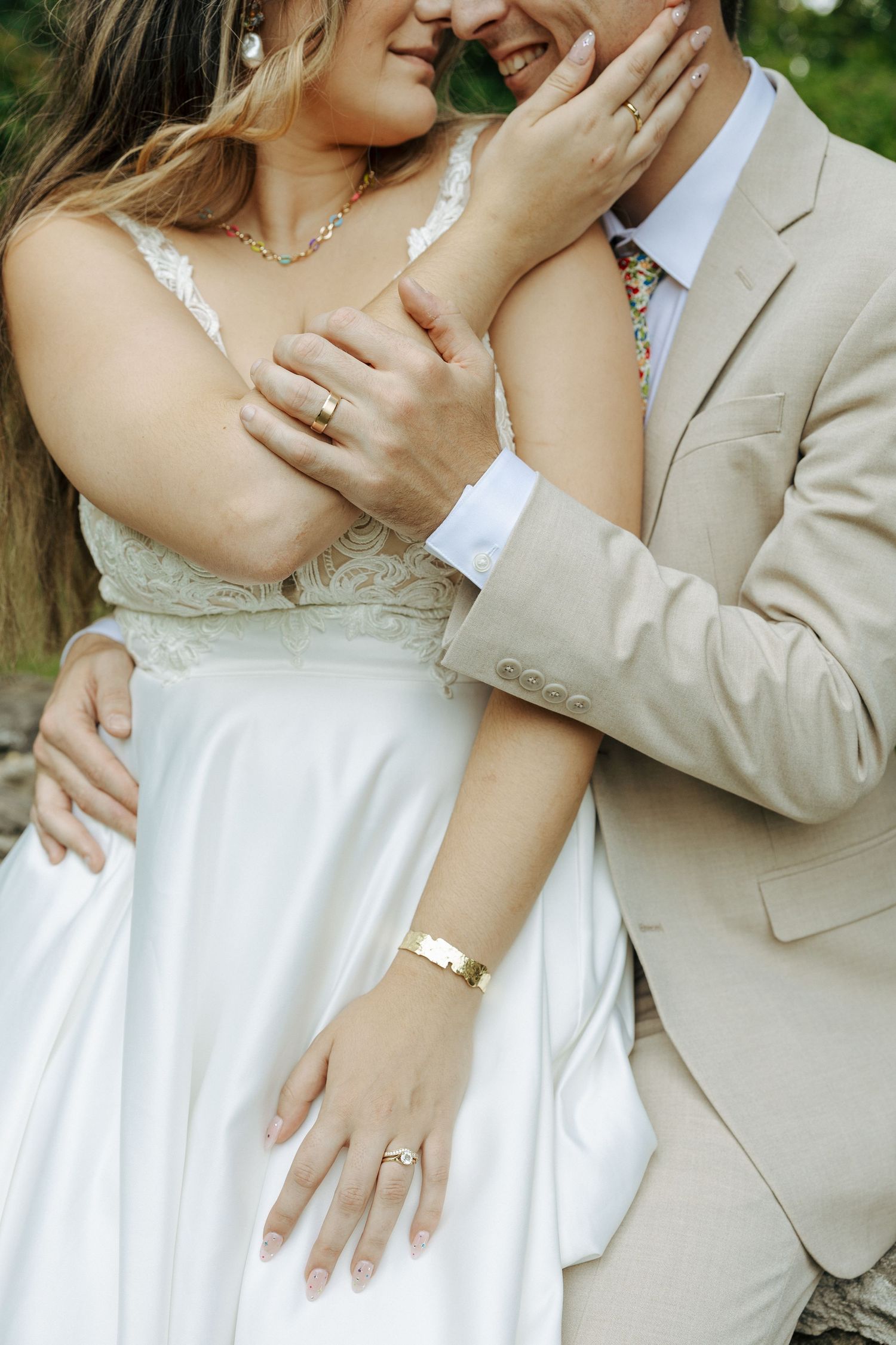Romantic close-up photo showing hands with wedding rings, white dress and beige suit details in outdoor garden setting.