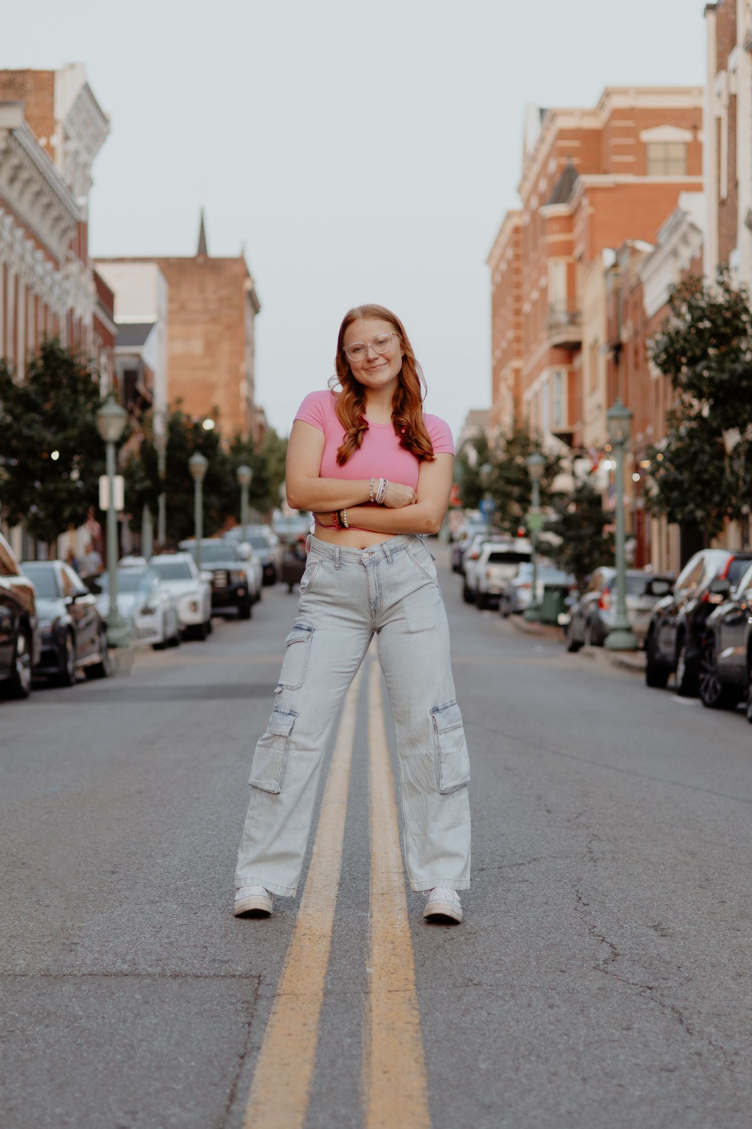 A woman in a pink top and light cargo pants stands on a city street with historic brick buildings and parked cars.