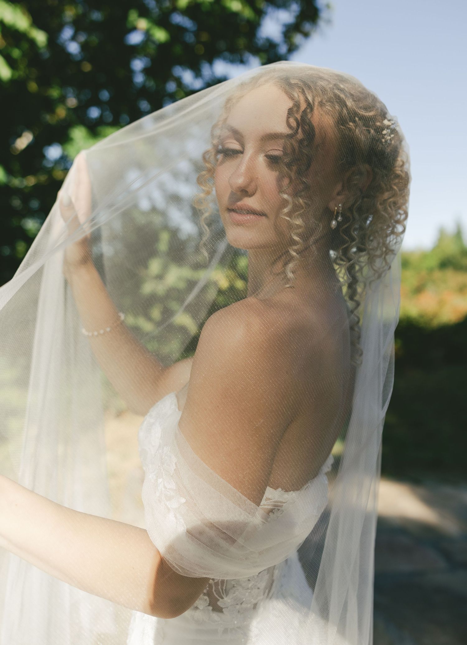 Ethereal bridal portrait outdoors showing delicate lace gown and flowing veil in soft natural lighting.