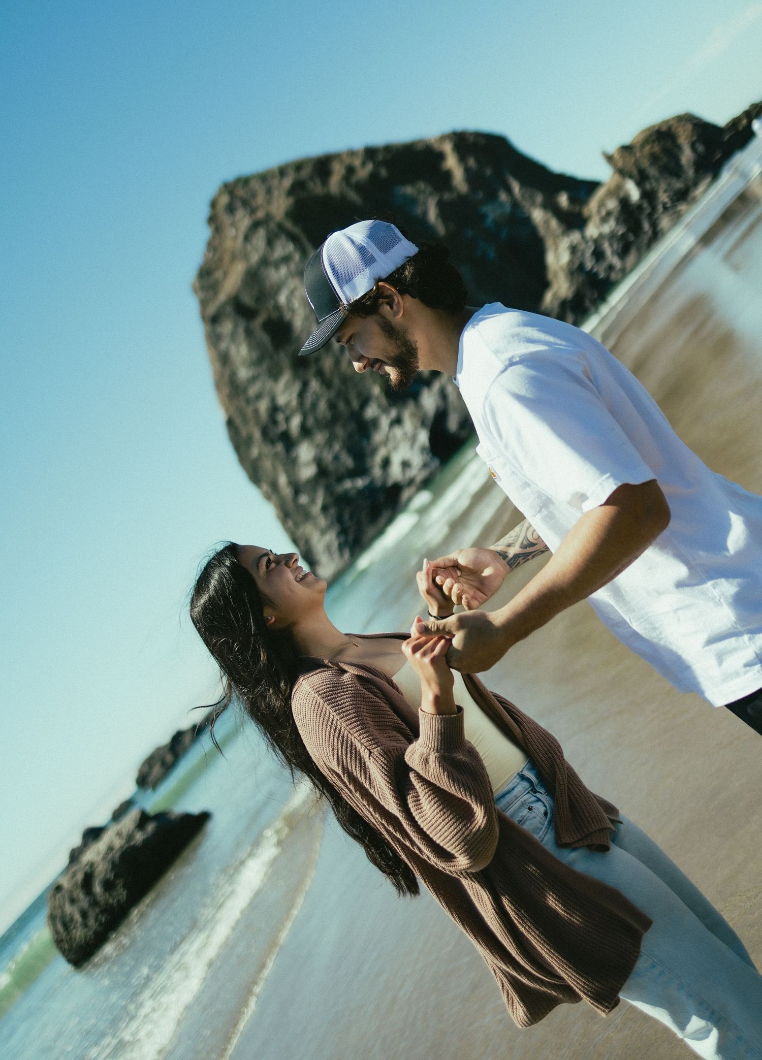 A couple shares a romantic moment on the beach with dramatic rocky cliffs in the background.