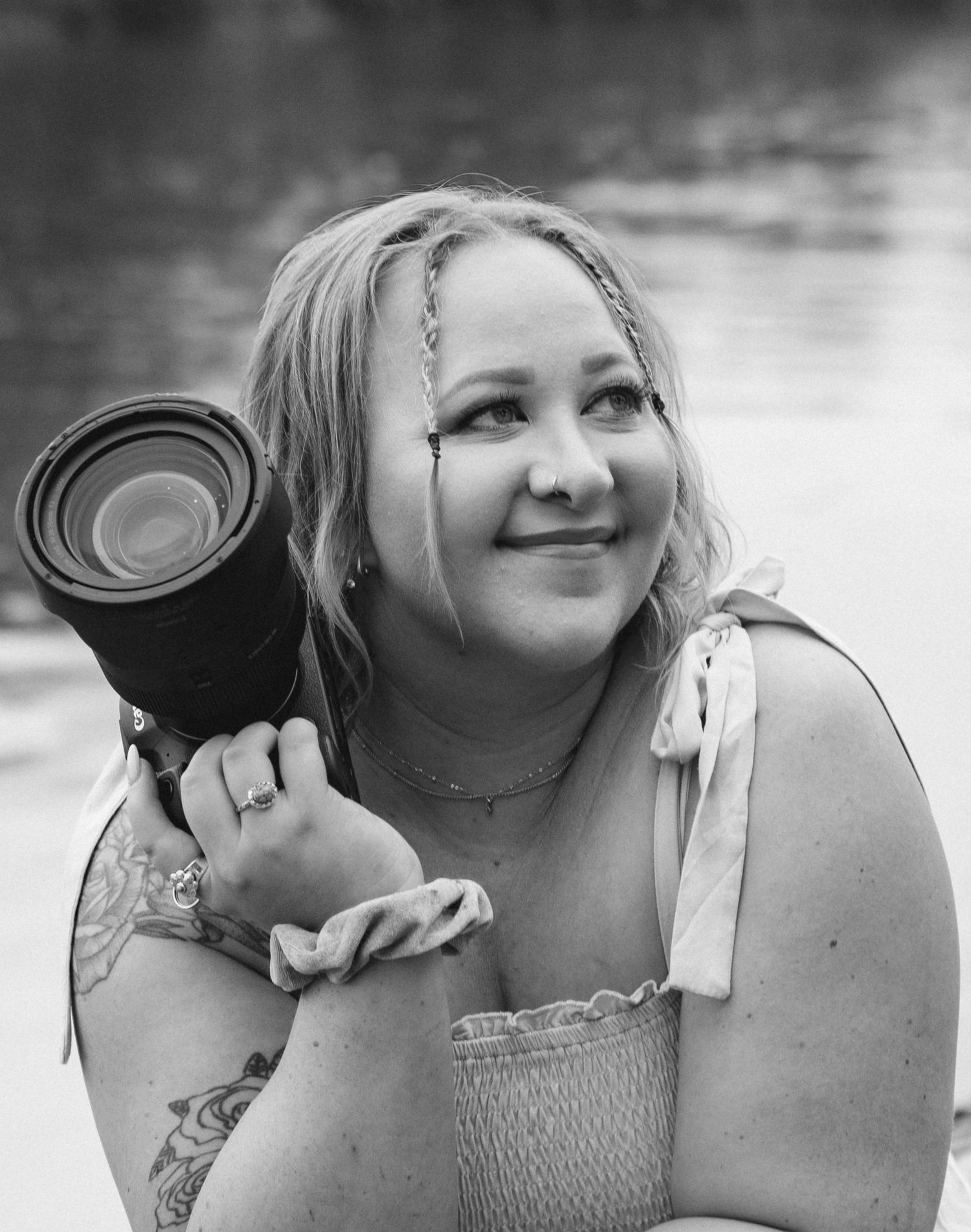 A photographer with braided hair smiles while holding a professional camera lens in a black and white portrait.