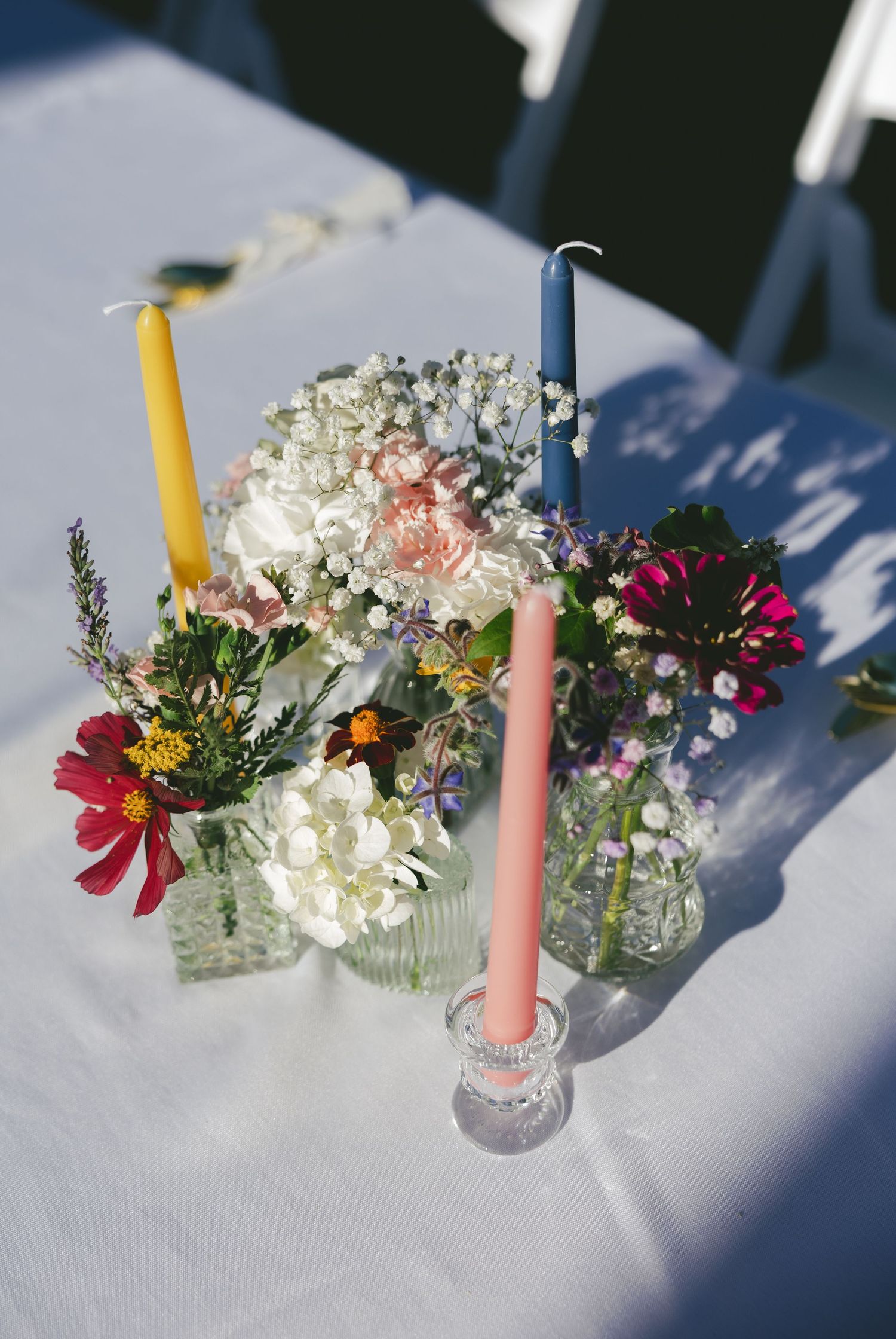 Row of small floral centerpieces with pink and white flowers in clear vases with pink and yellow candles on white tablecloth.