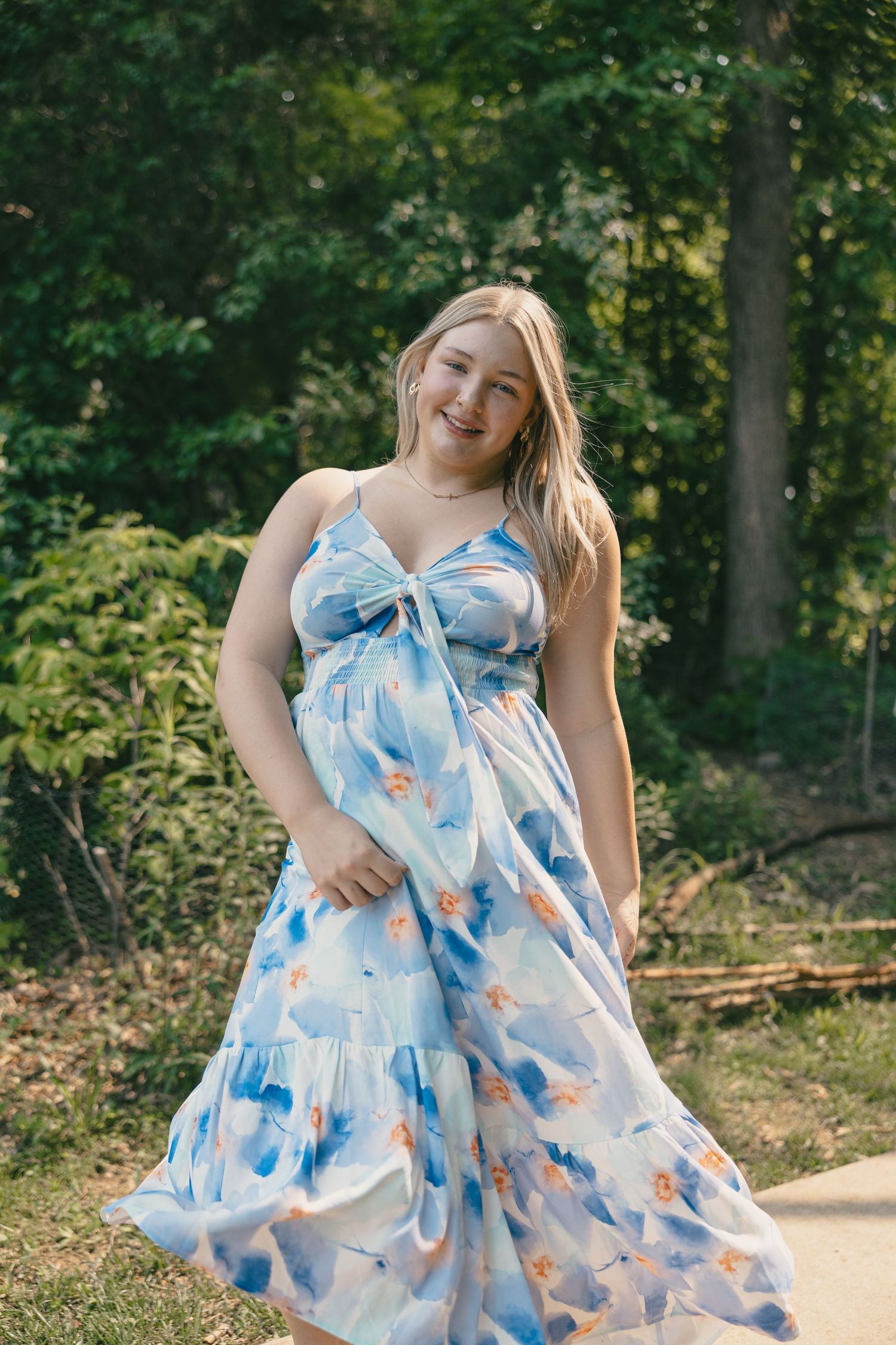 Someone in a flowing blue and white floral maxi dress twirls on a path surrounded by green trees in a park setting.