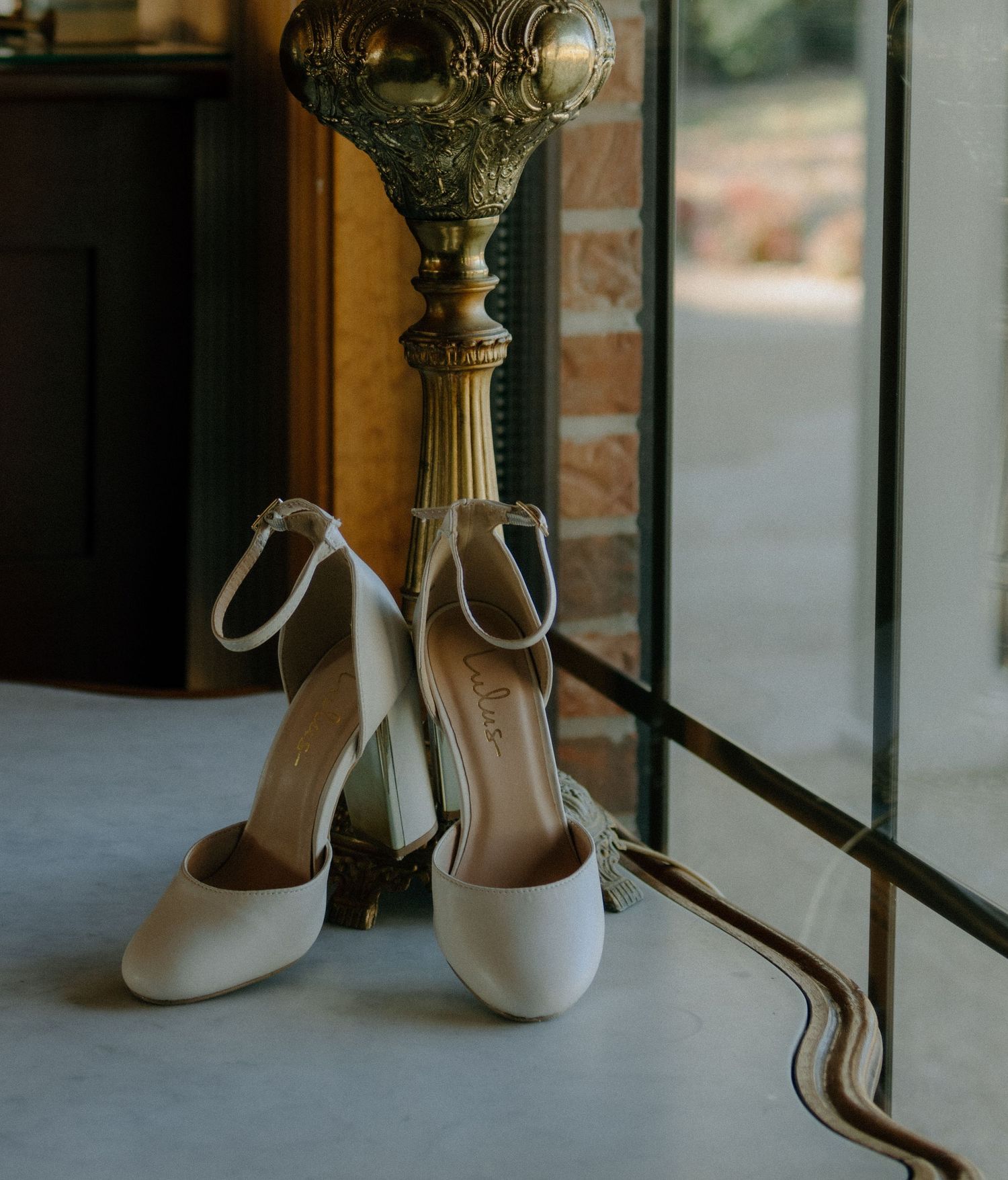 Elegant white bridal heels placed next to an ornate vintage lamp stand by a window.