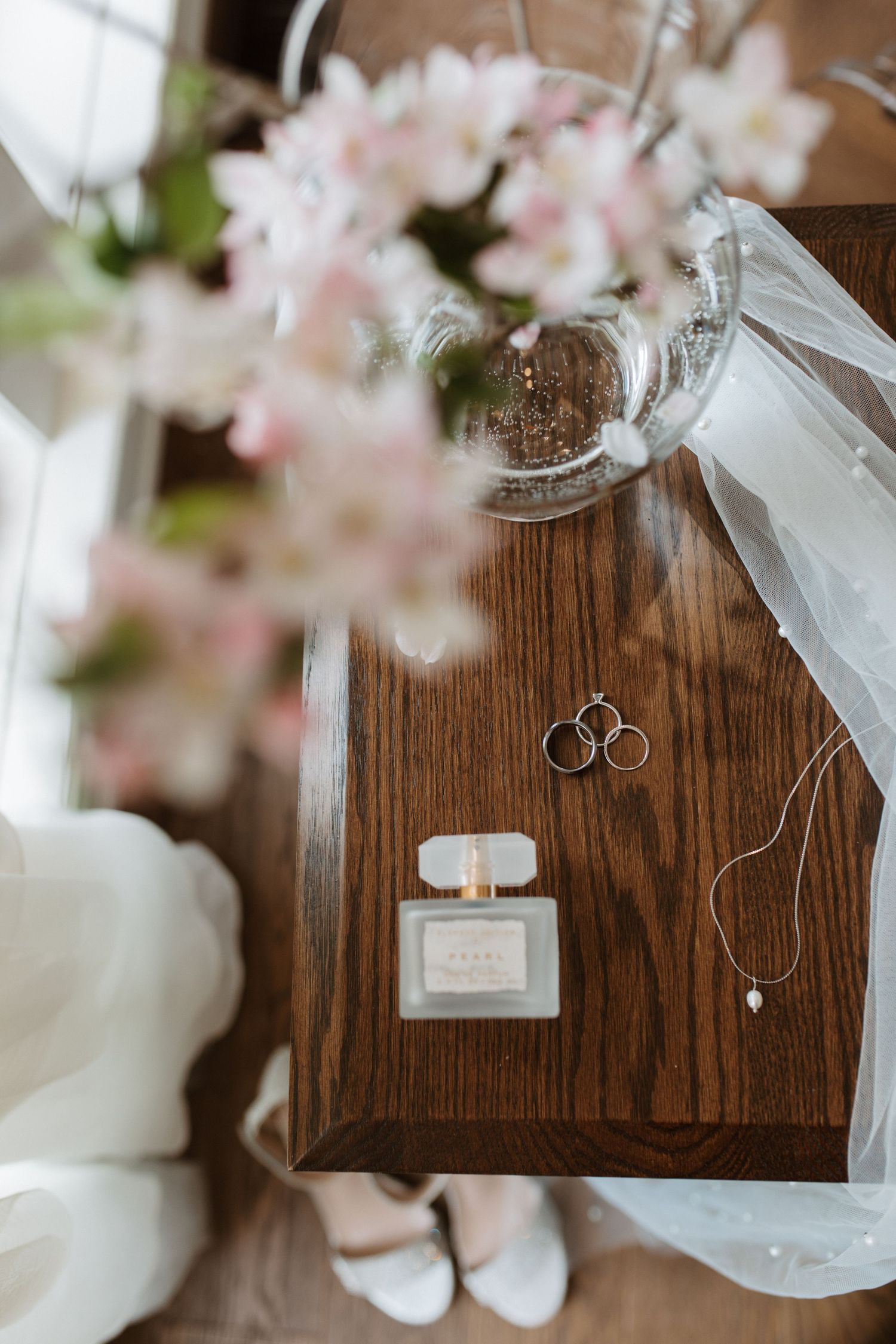Wedding accessories including rings, perfume bottle, and white shoes arranged on wooden surface with soft pink flowers.