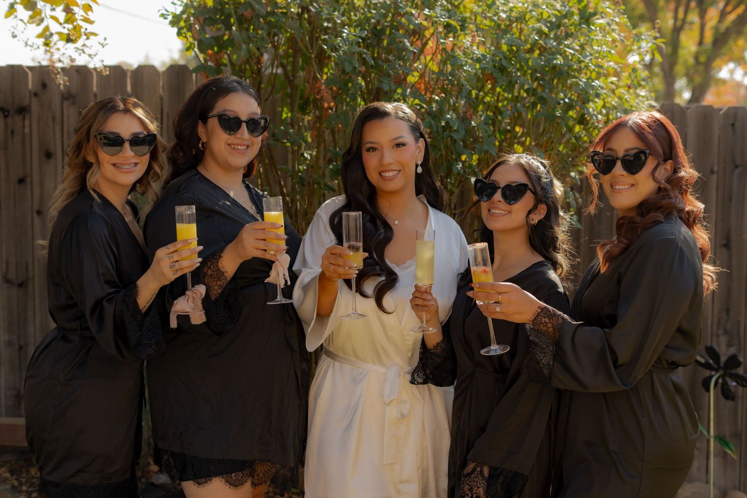 Group of friends in dark attire toasting with drinks in a sunny garden setting.