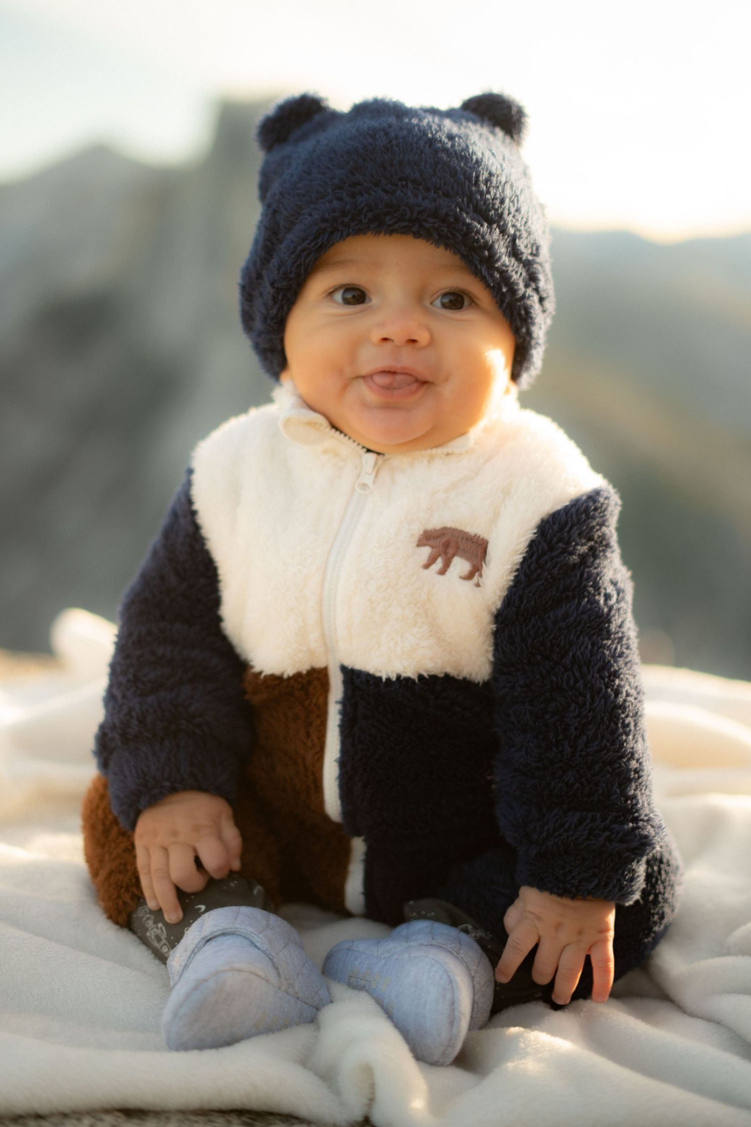 Baby in bear-themed winter outfit and beanie sits on blanket with mountains in background.