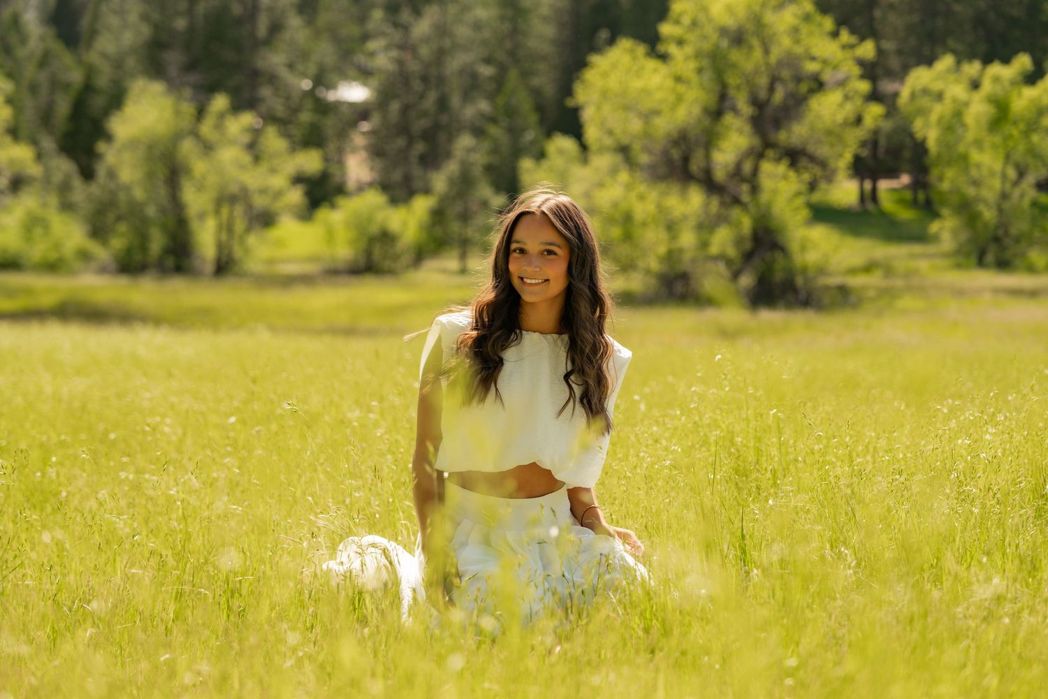 Woman in white outfit sitting in lush green meadow with trees, smiling outdoors in natural sunlight.