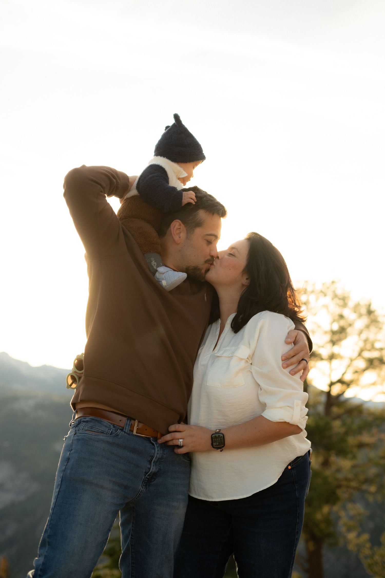 Family portrait at sunset with baby perched on shoulders as parents share romantic moment.