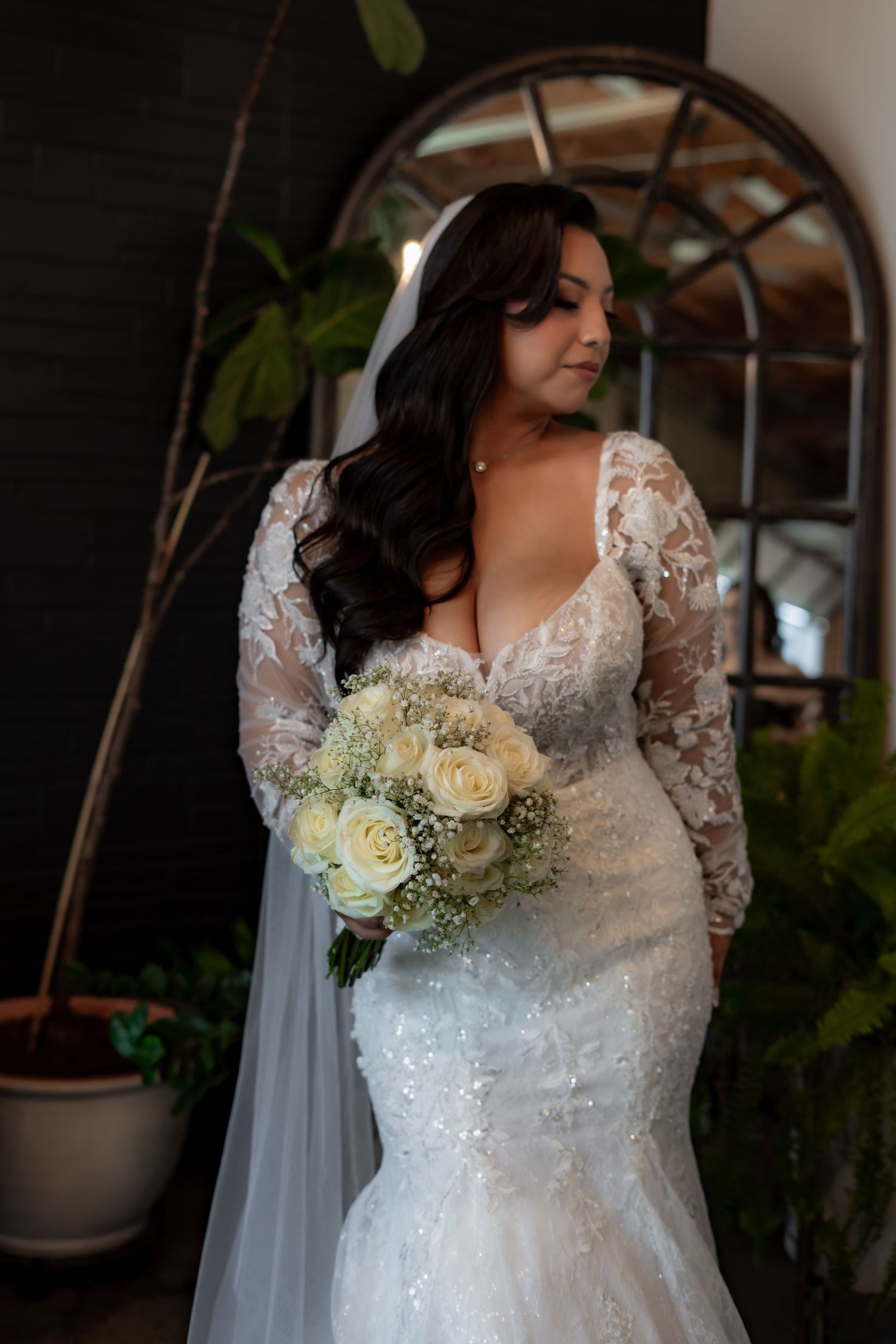 A bride in a lace white wedding dress holds an elegant bouquet of cream roses and greenery against an indoor setting.