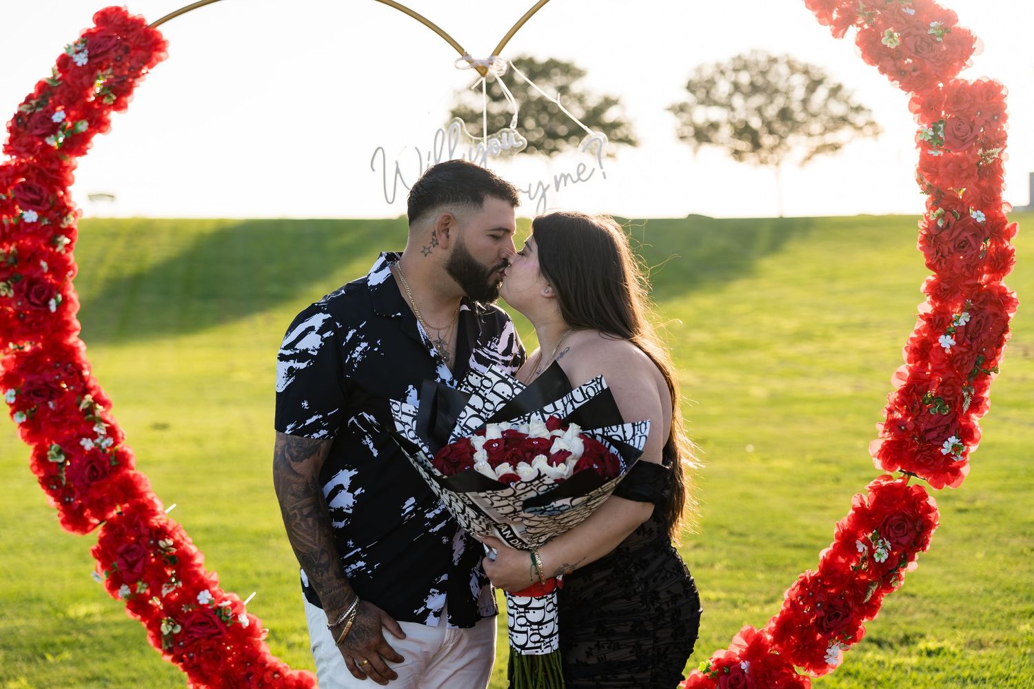 A couple poses against a heart-shaped red floral arch in an outdoor garden setting at sunset.