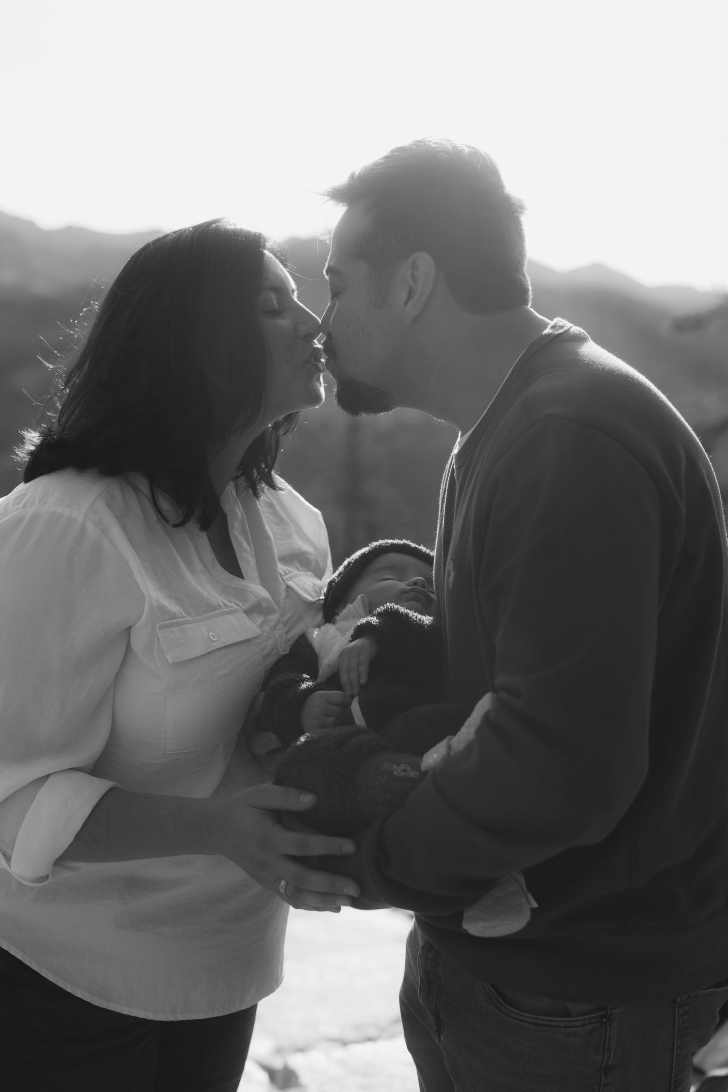 Intimate moment between couple sharing affection while holding their baby outdoors.