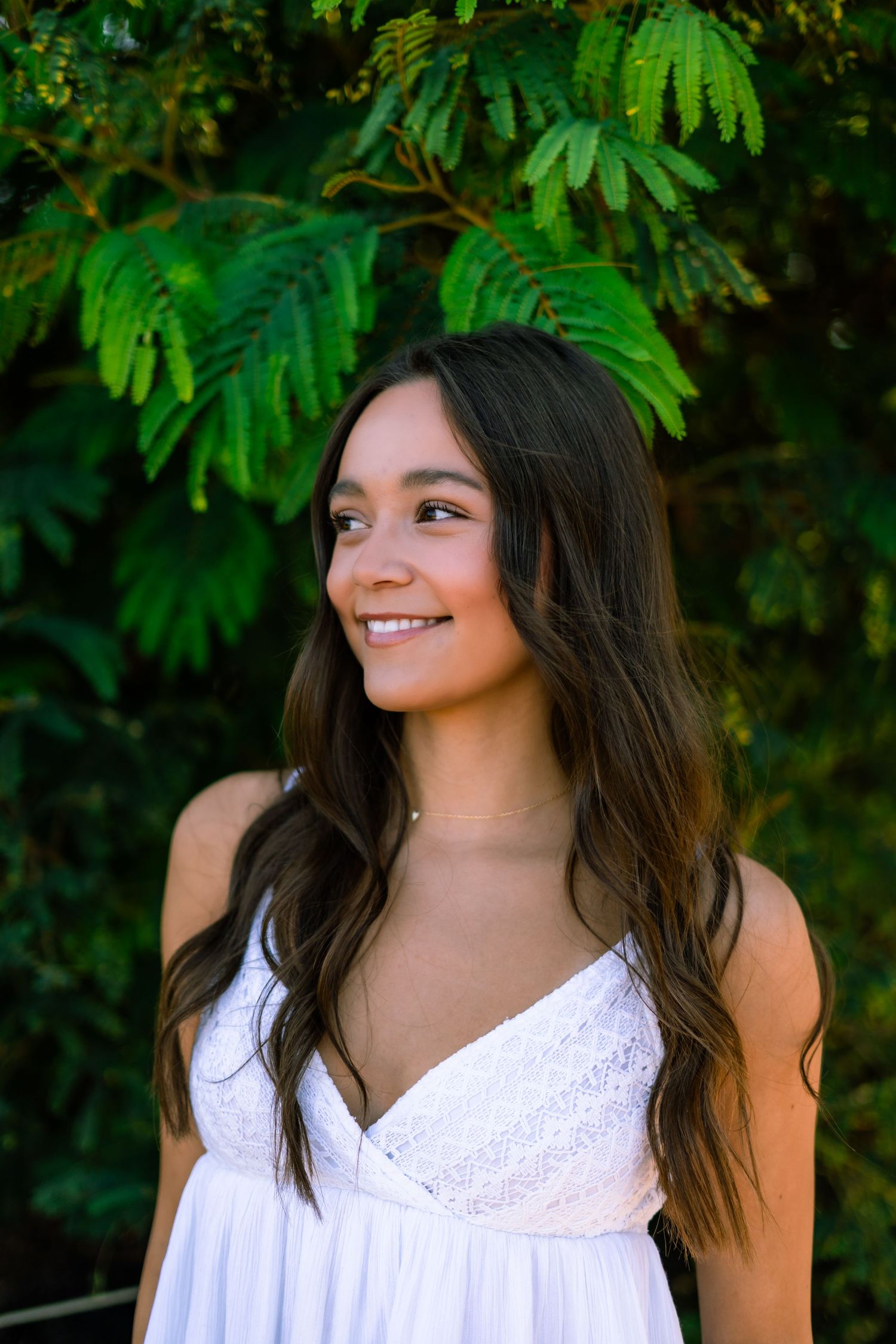 A smiling person in a white sundress poses outdoors against lush green foliage in a natural setting.